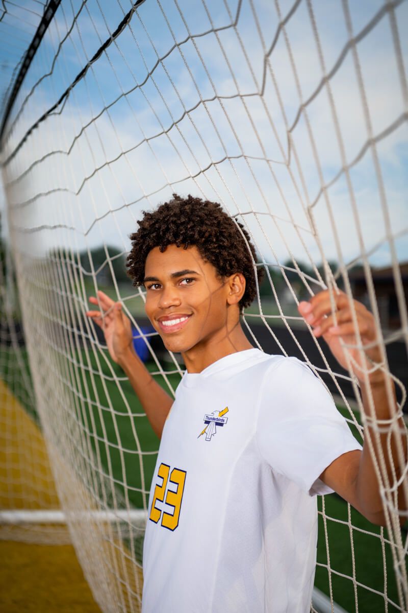 Smiling teen soccer player in white #23 jersey poses behind goal net on a sunny day.