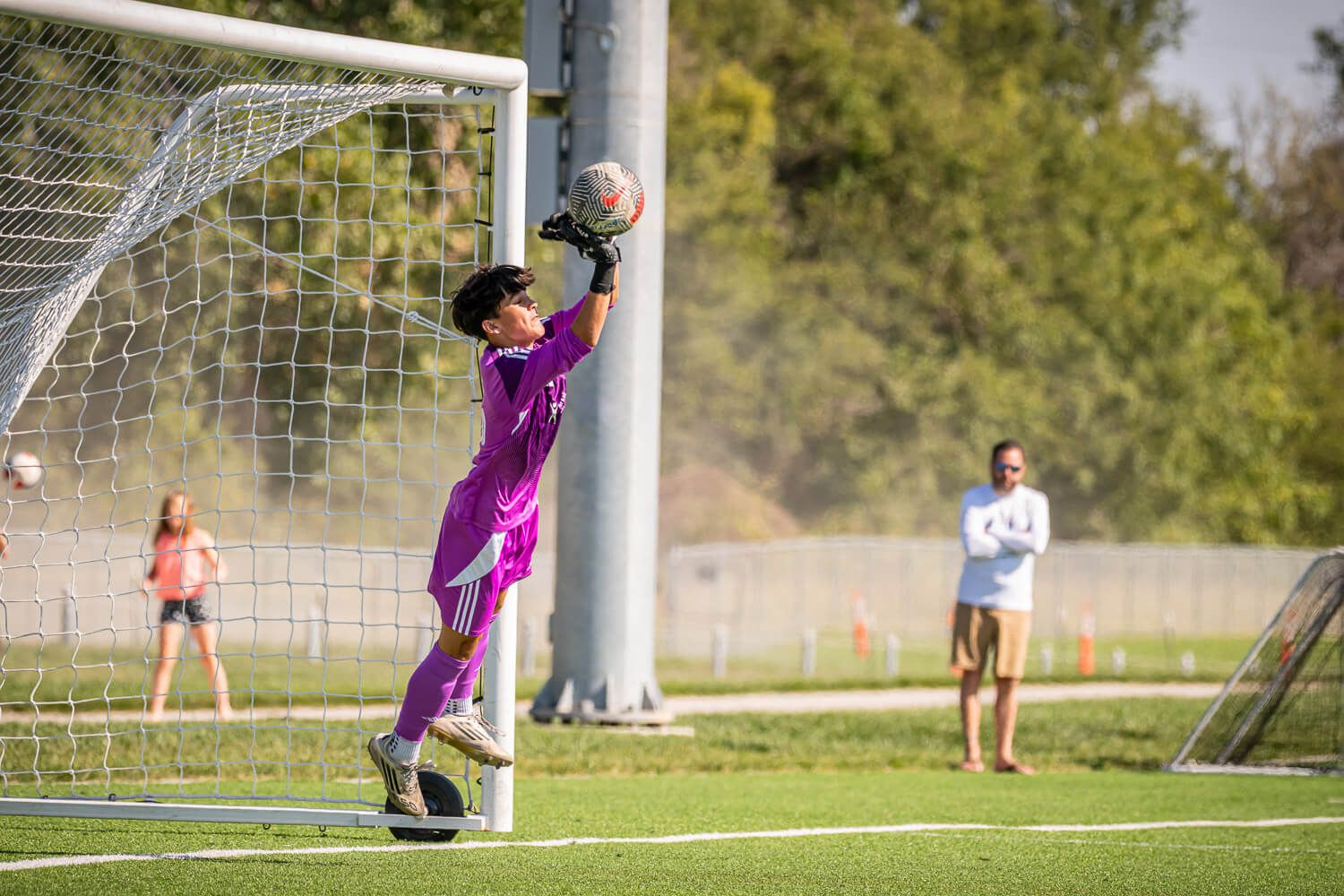 Soccer goalkeeper in purple jersey makes diving save during outdoor match on sunny day.