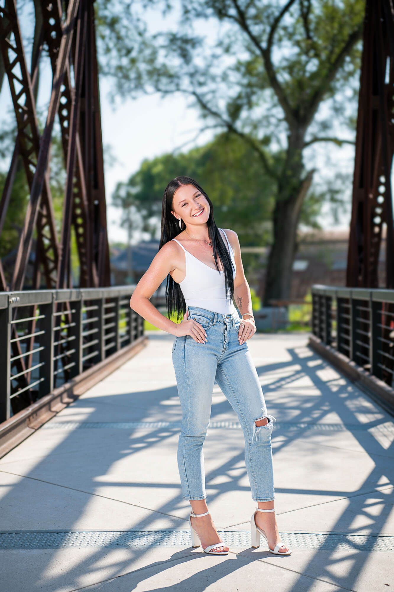 A woman poses on a metal bridge wearing ripped jeans, white top and heels on a sunny day.