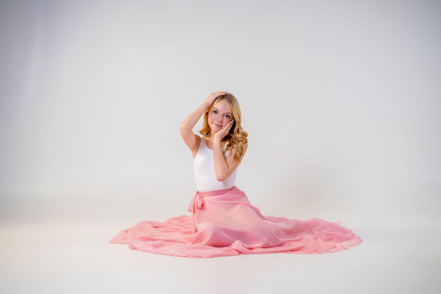 Omaha Senior Photography - Photographer in Omaha - Figure in white top and flowing coral pink skirt sits gracefully on studio floor.