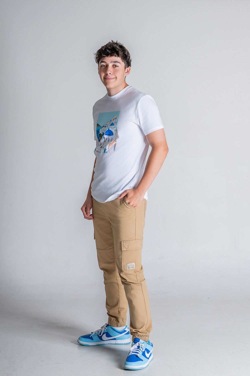 Teen boy in white graphic tee, tan cargo pants, and blue Nike sneakers posing against a gray background.