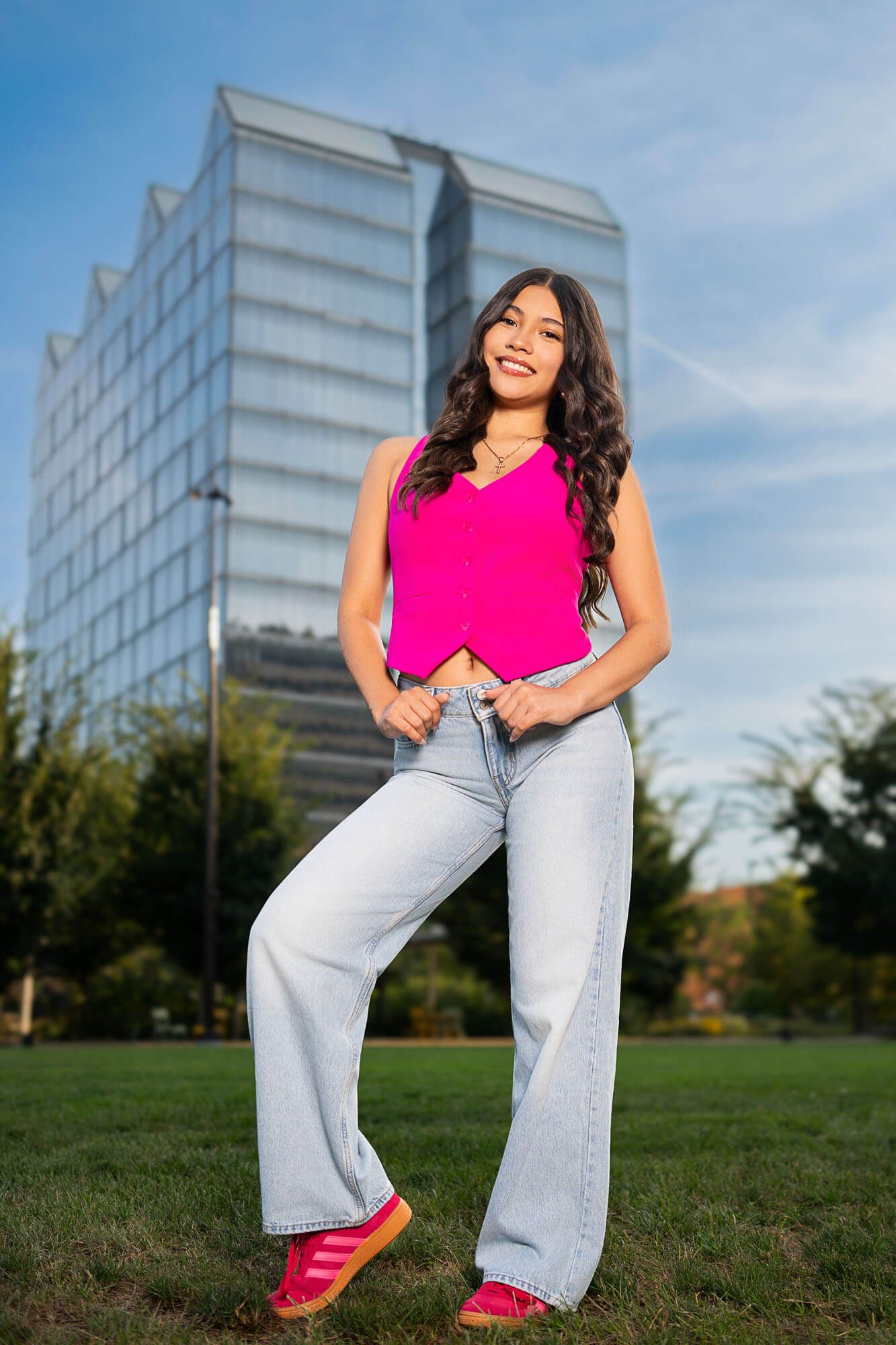 A vibrant pink crop top and light jeans outfit photographed against a modern glass building and green lawn.
