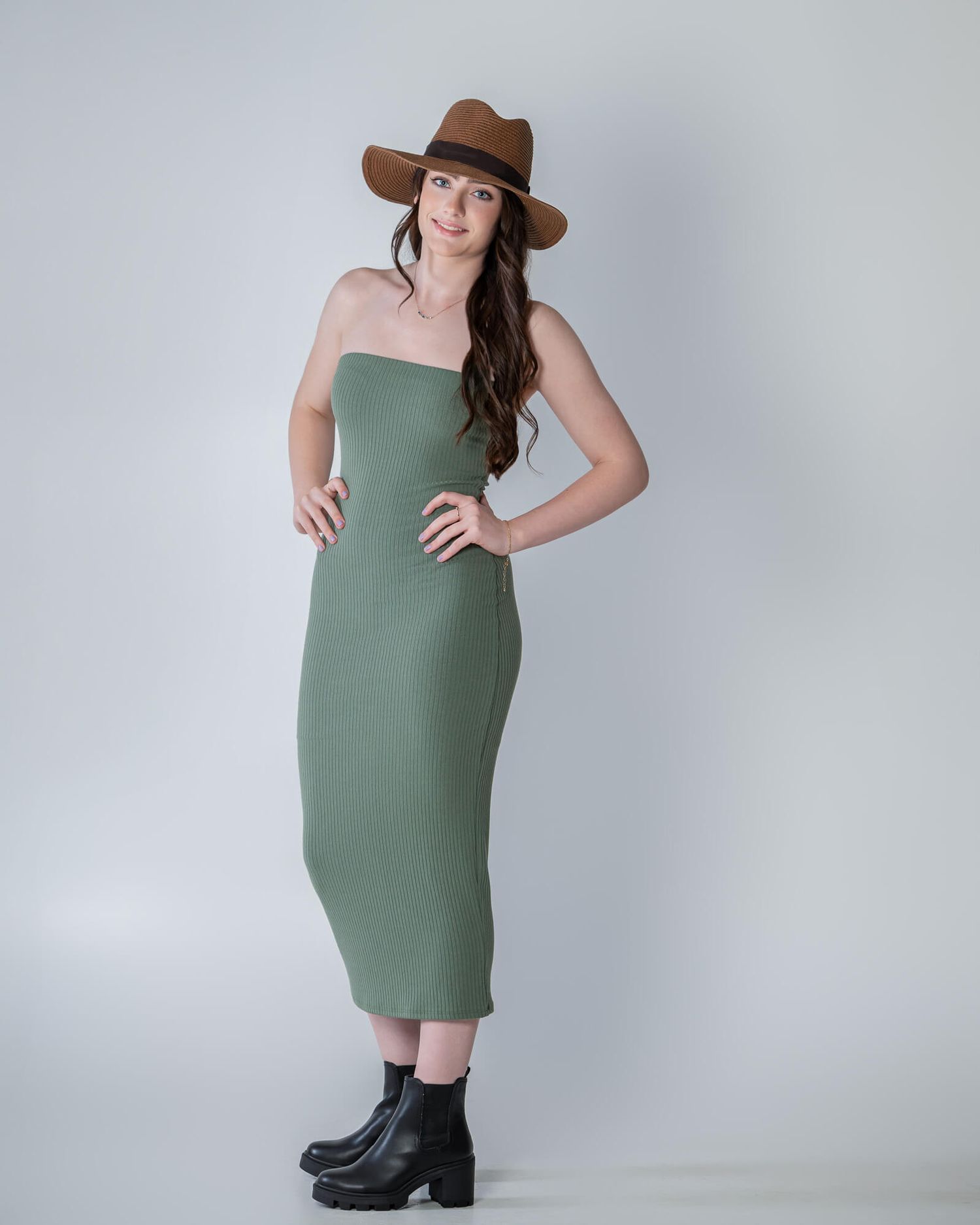 Woman in green dress and brown hat poses with black boots in studio against white backdrop.