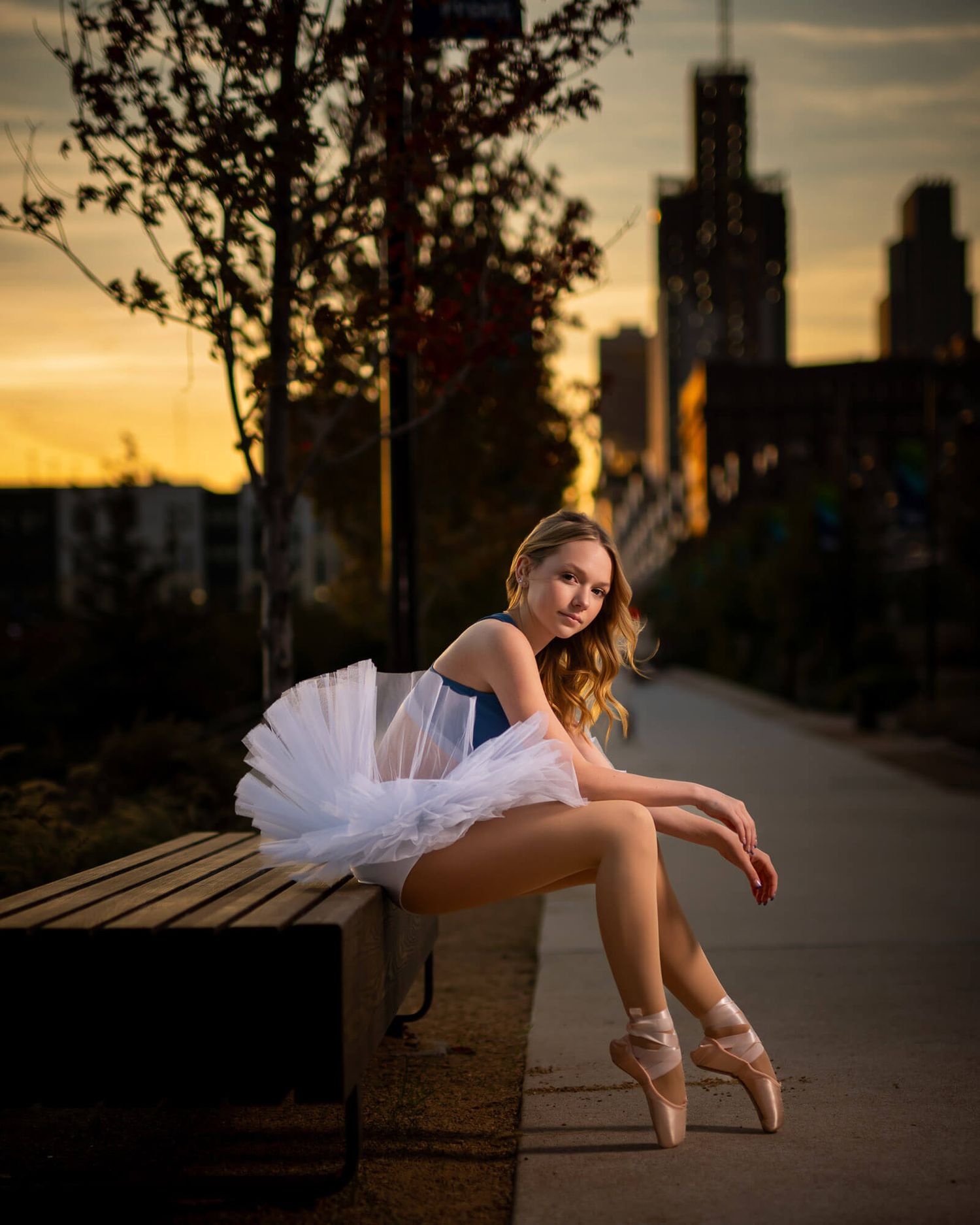 High school senior picture of ballet dancer in white tutu sits gracefully on bench at sunset with Omaha skyline silhouetted in background.
