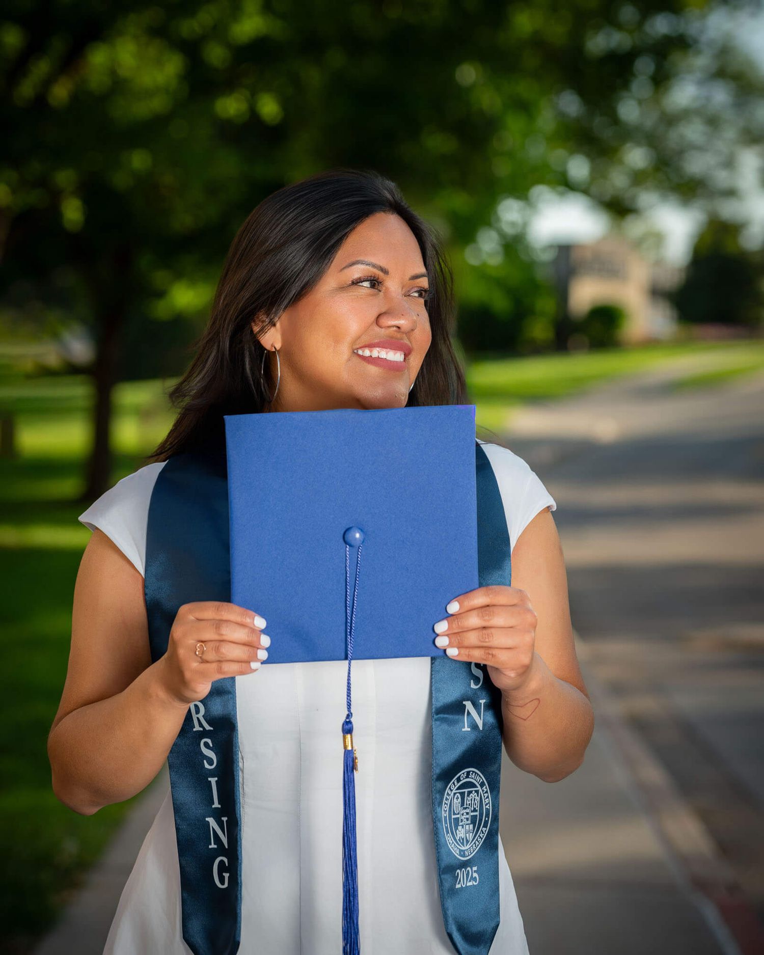 A smiling nursing graduate holds her blue mortarboard cap outdoors, wearing a white dress and nursing graduation sash.