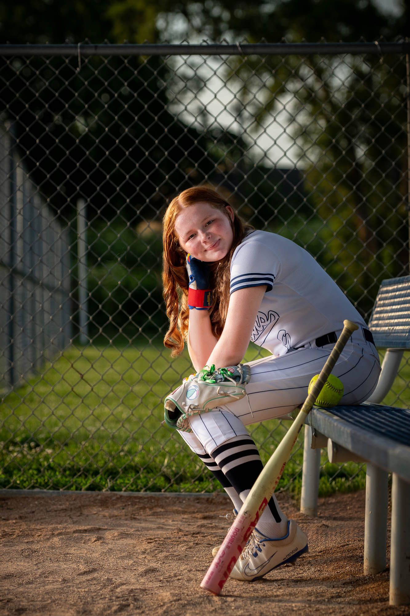 Young female softball player in uniform sits on dugout bench holding bat, smiling at camera near chain-link fence.