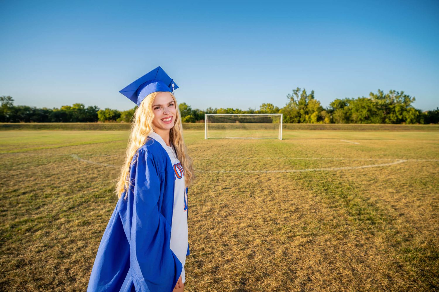 Smiling blonde graduate in blue cap and gown stands on soccer field with goal post visible in background.
