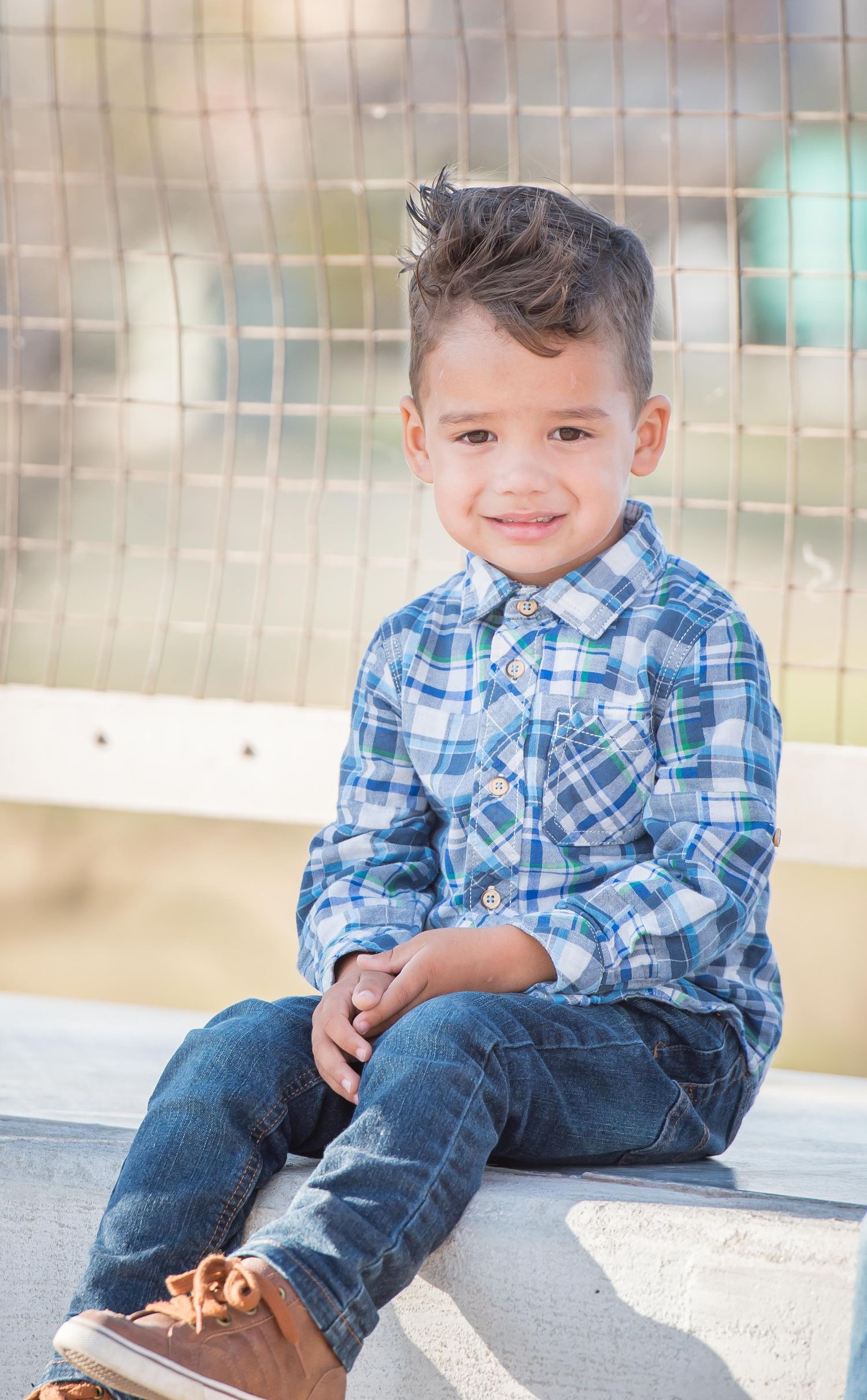 Young boy wearing a blue plaid shirt and jeans sits casually on a bench with a happy expression.