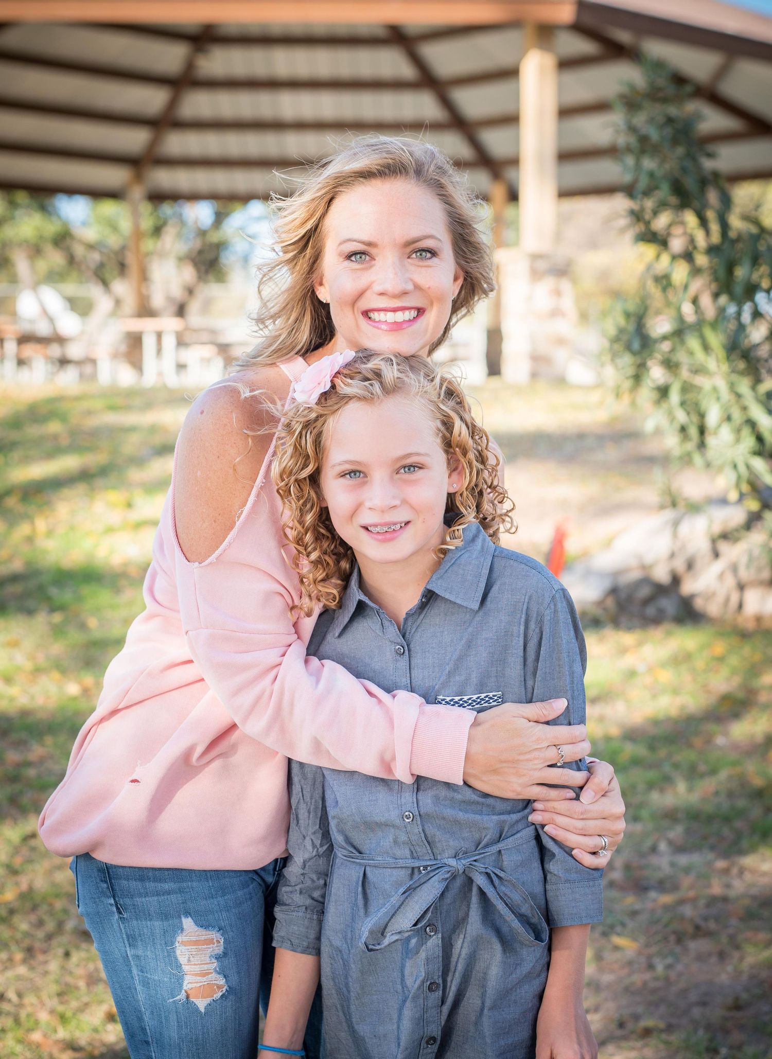 Mom and daughter smile warmly while embracing under a wooden gazebo structure in a sunny outdoor setting during family photos in Omaha.