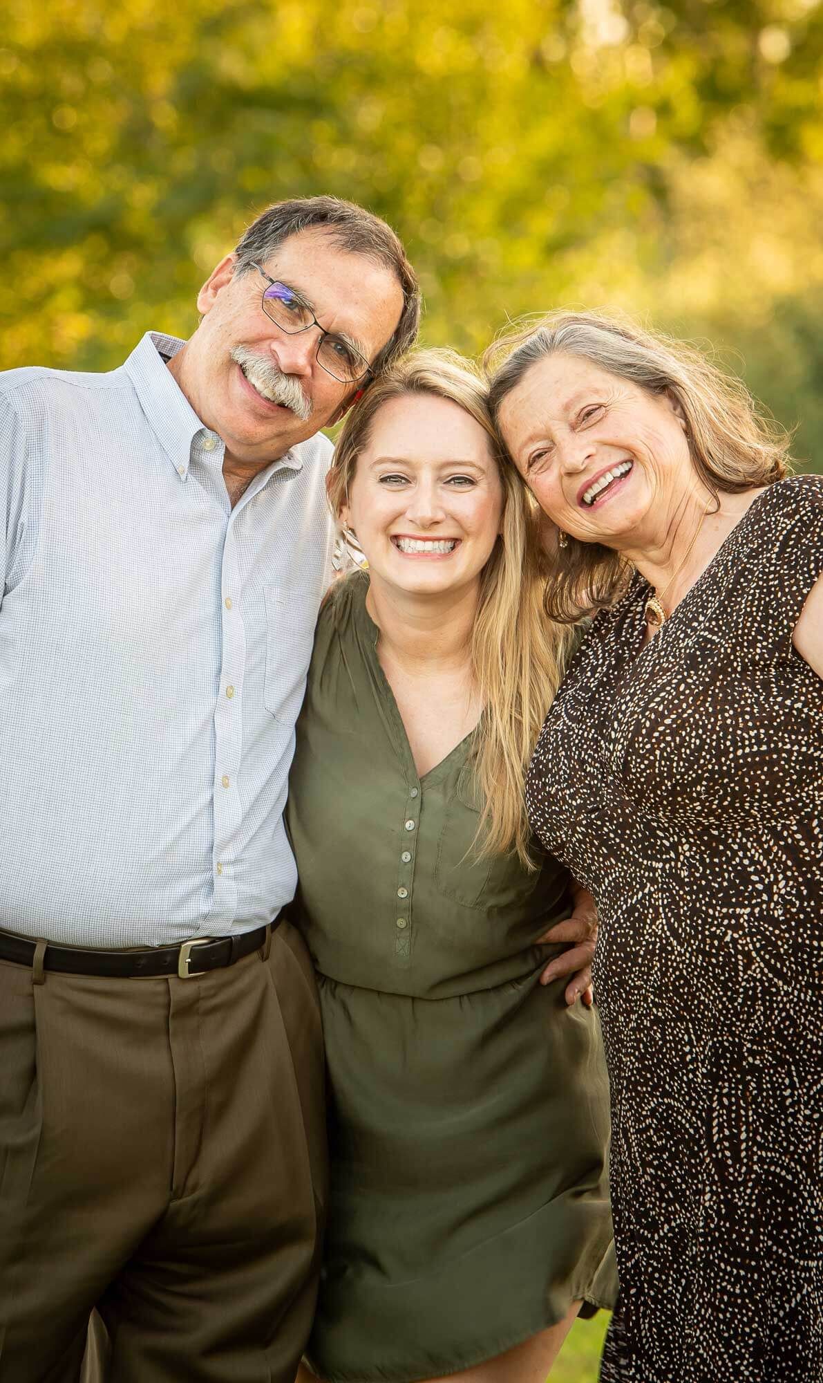 Three people embrace and smile warmly while posing together in a sunny outdoor setting.