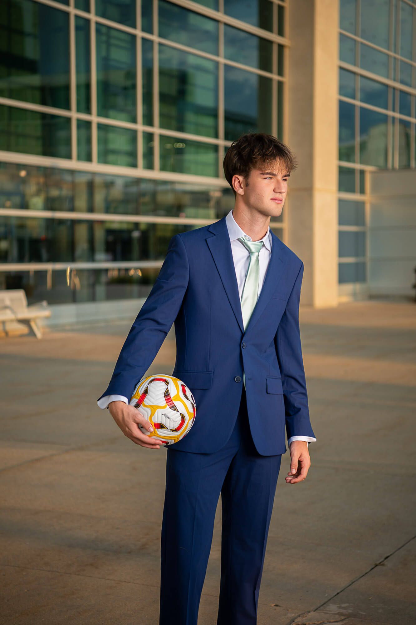 Professional in a navy suit holds a volleyball while posing outside a modern glass building.