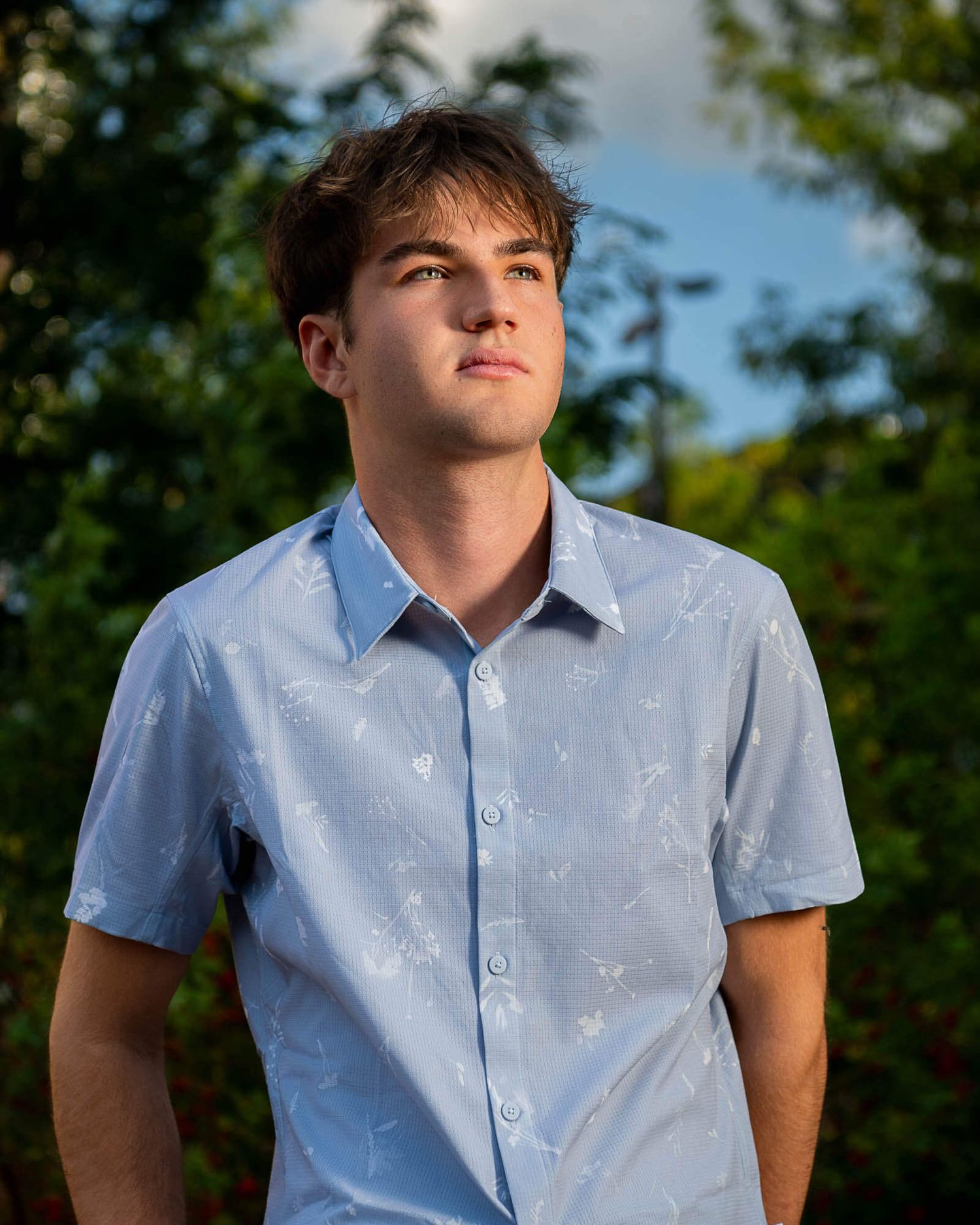 Person in light blue short-sleeve button down shirt poses outdoors during golden hour lighting.