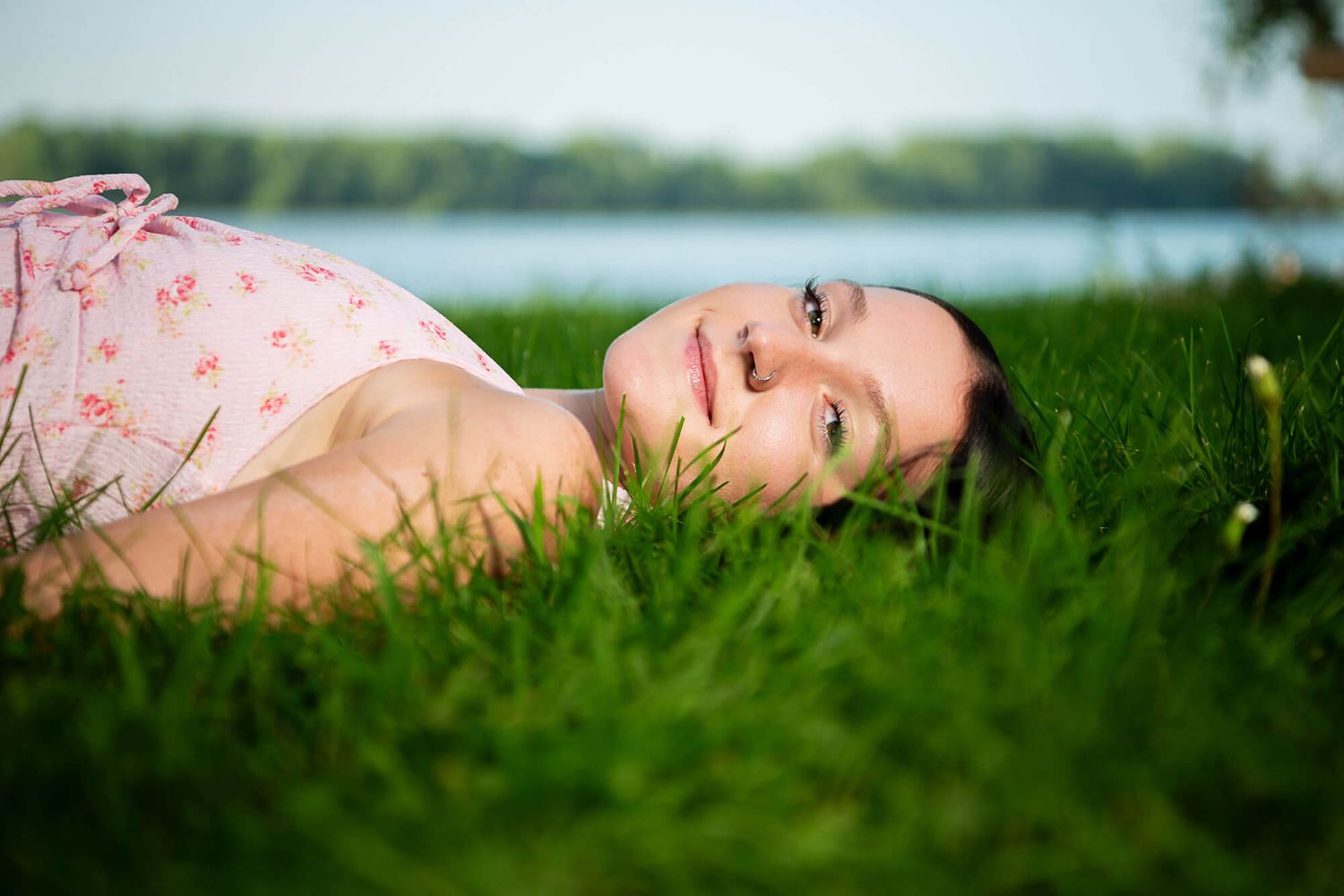 Person lying in lush green grass near a tranquil lake during a sunny day.