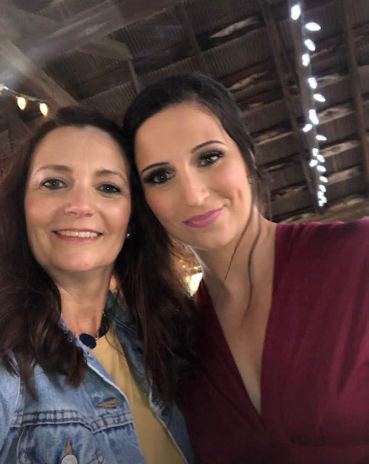 Two women taking a selfie together inside a rustic venue with string lights visible in the background.