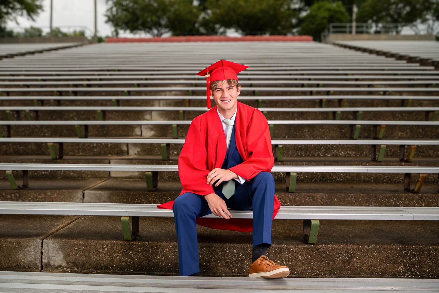 Graduate in red cap and gown sits on concrete stadium bleachers during outdoor photo session.