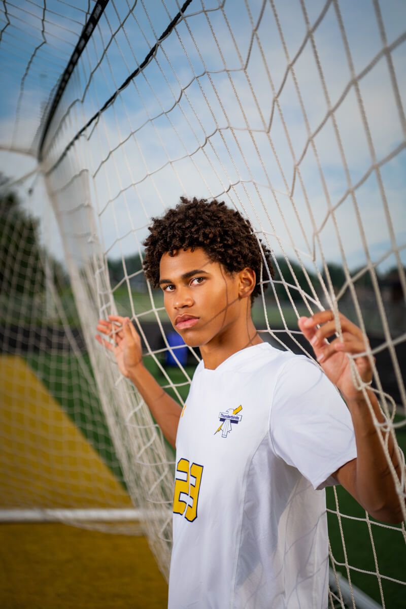 Young male soccer player in white #23 jersey stands pensively behind a goal net outdoors.