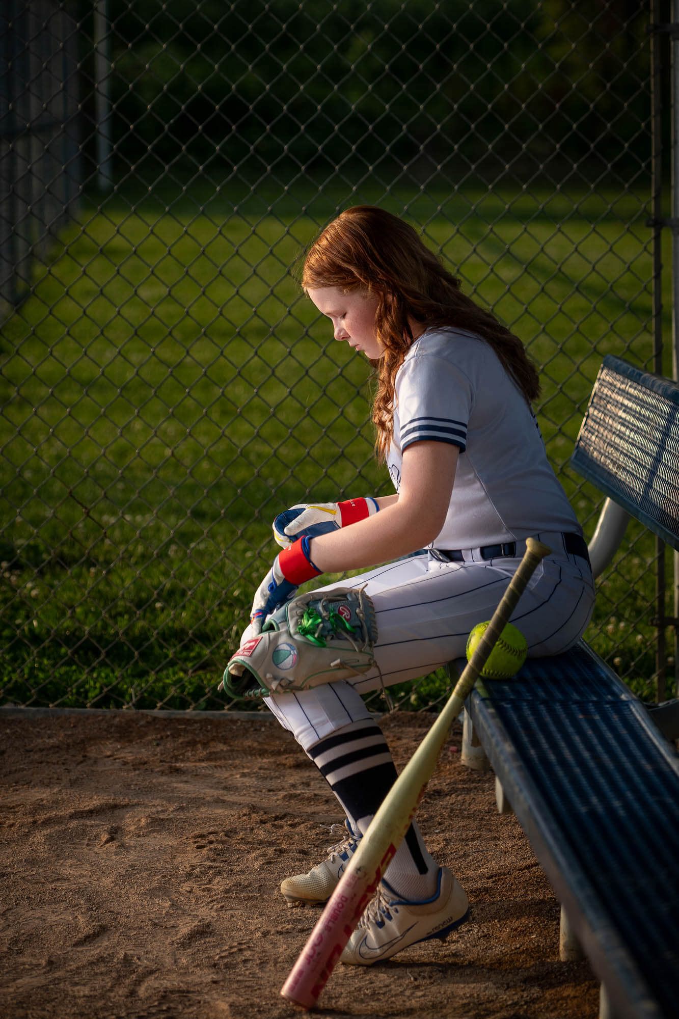 Young female softball player in uniform sitting on dugout bench with bat, wearing batting gloves at baseball field.