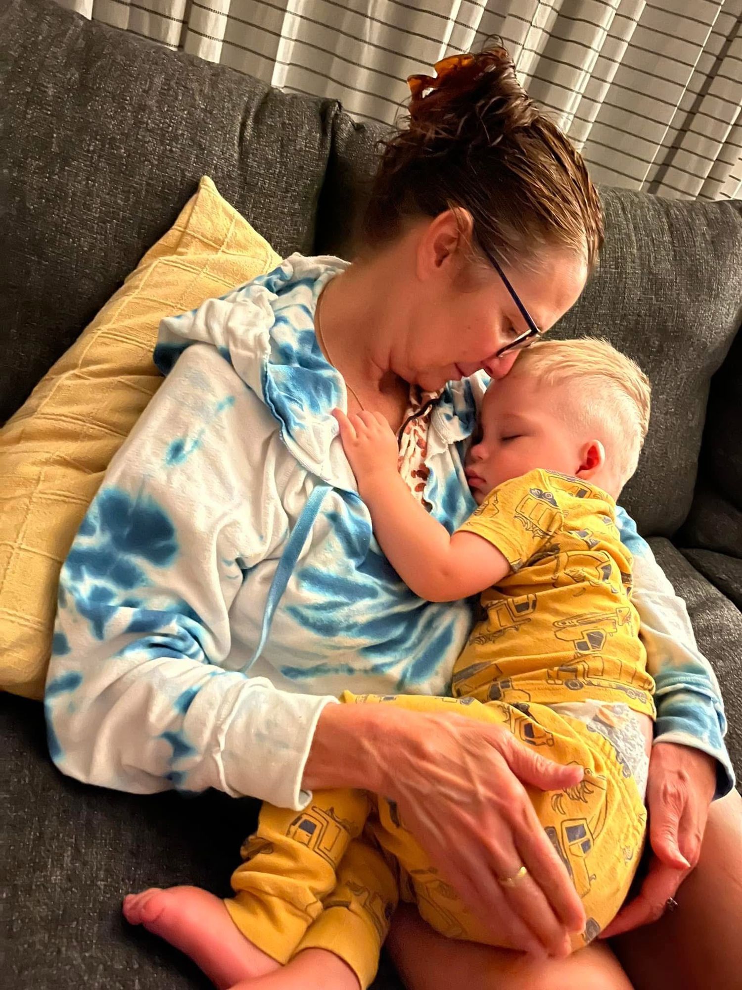 A parent in a tie-dye sweatshirt cuddles with a baby wearing yellow pajamas while resting together on a gray couch.