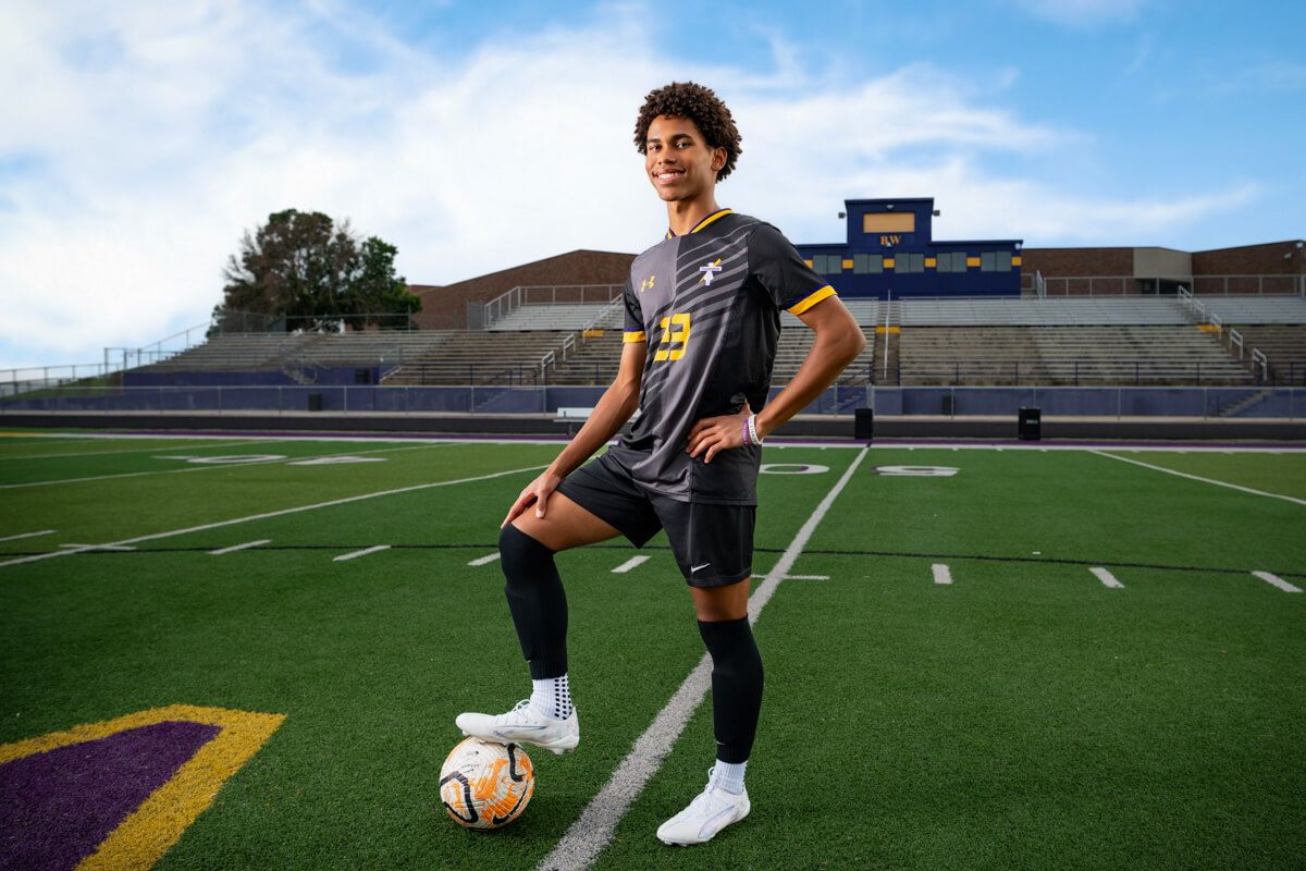 A soccer player practices dribbling drills on an artificial turf field with goal posts visible in the background.