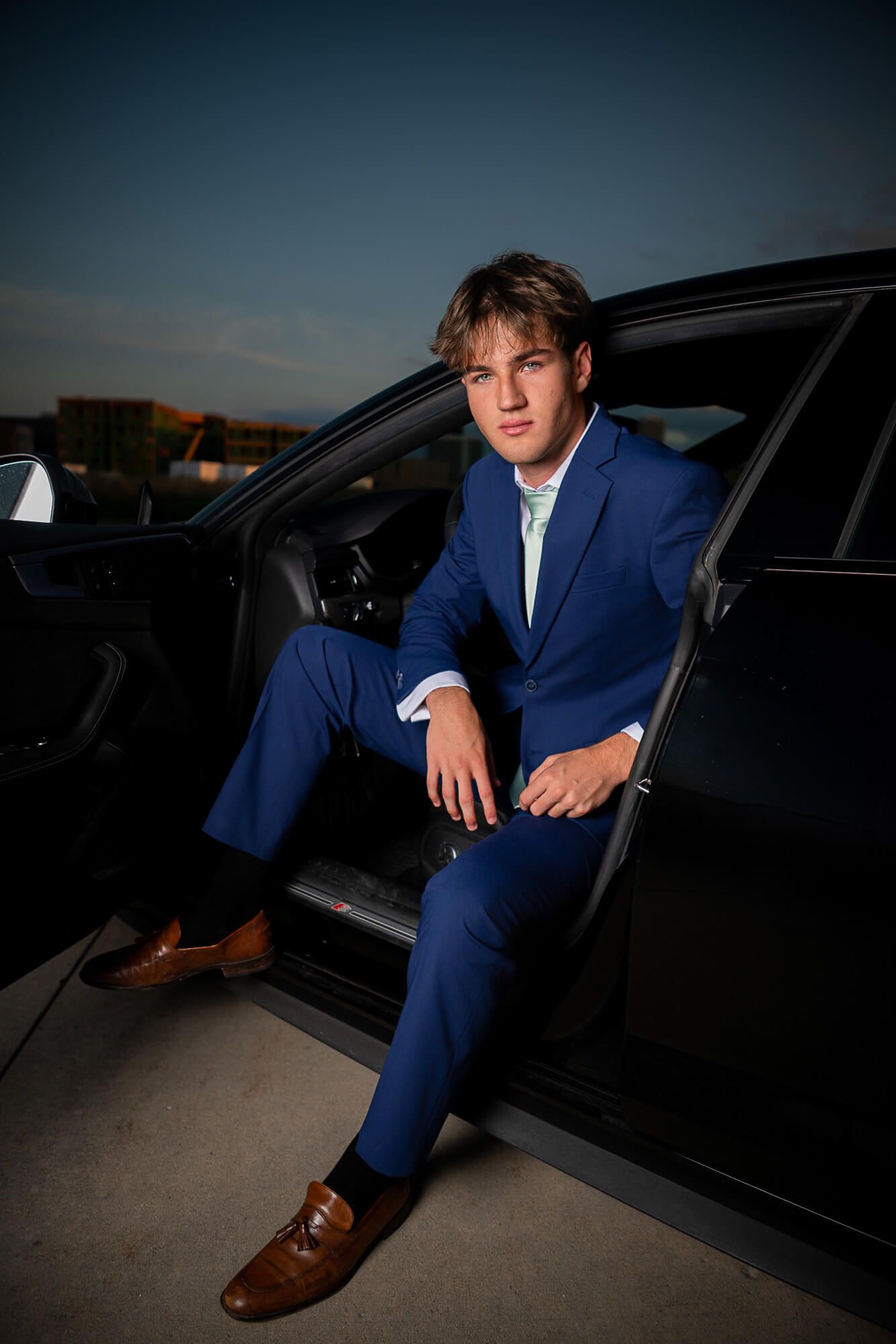 Graduate from Westside High School during senior photography session sits in his Audi with sunset in the background wearing a blue suit.