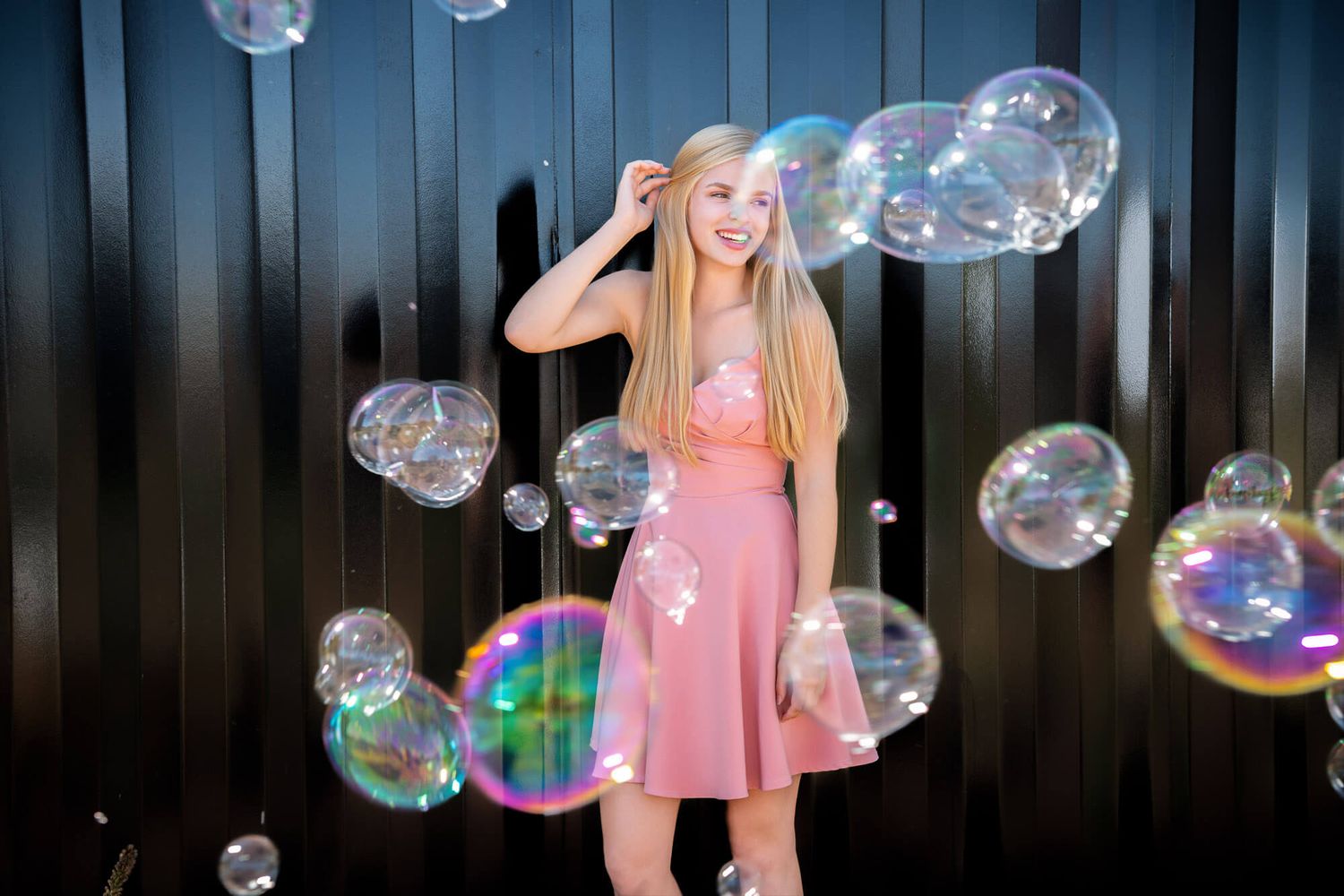 Young blonde girl in pink dress poses among large colorful soap bubbles in front of dark metal wall.