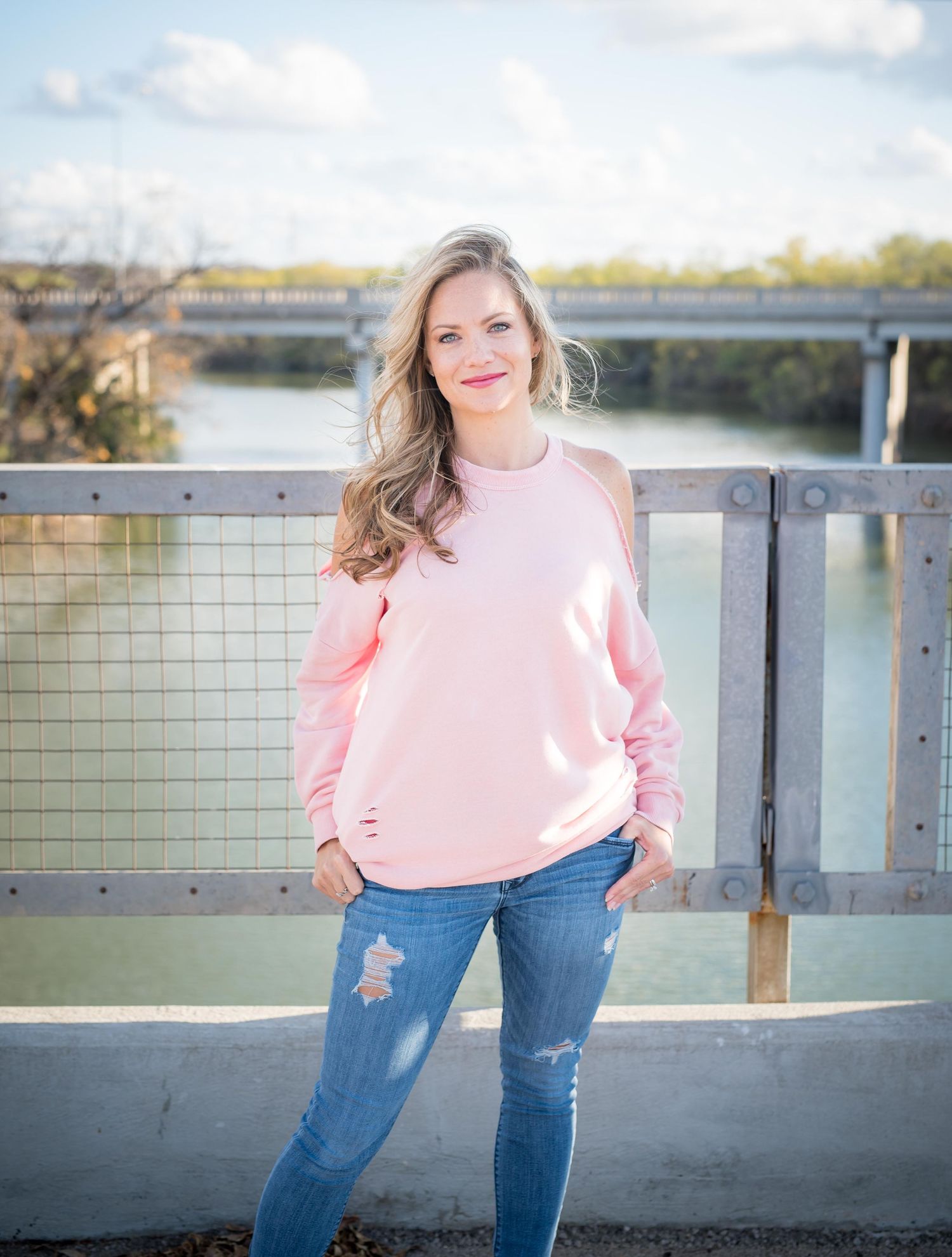 Person wearing a pink sweater and distressed jeans poses on bridge railing with river and cloudy sky in background.