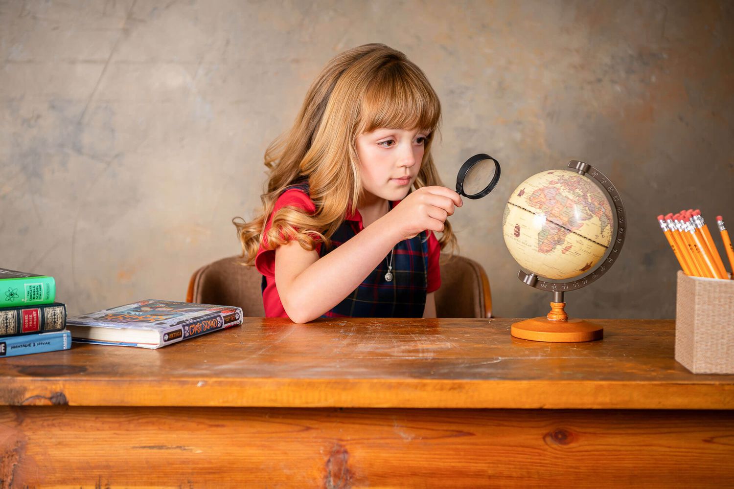 A student sits at a wooden desk with various school supplies and a globe, examining items with a magnifying glass.