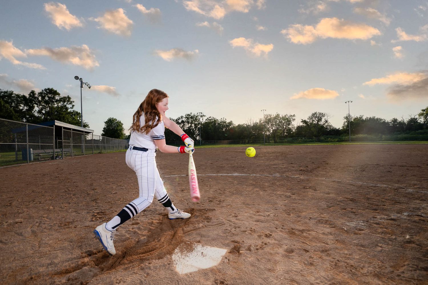 Young female softball player swinging a pink bat at a yellow ball on a dirt field at sunset.