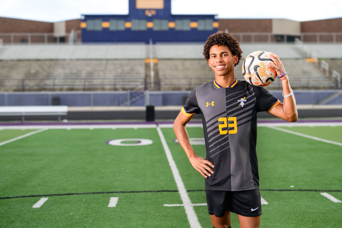 Young soccer player in black Under Armour jersey number 23 poses with ball on green turf field at stadium.