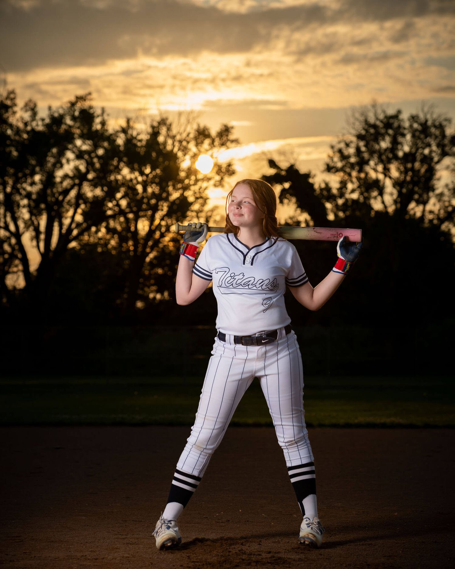 Softball player poses with bat at sunset wearing white uniform on field.