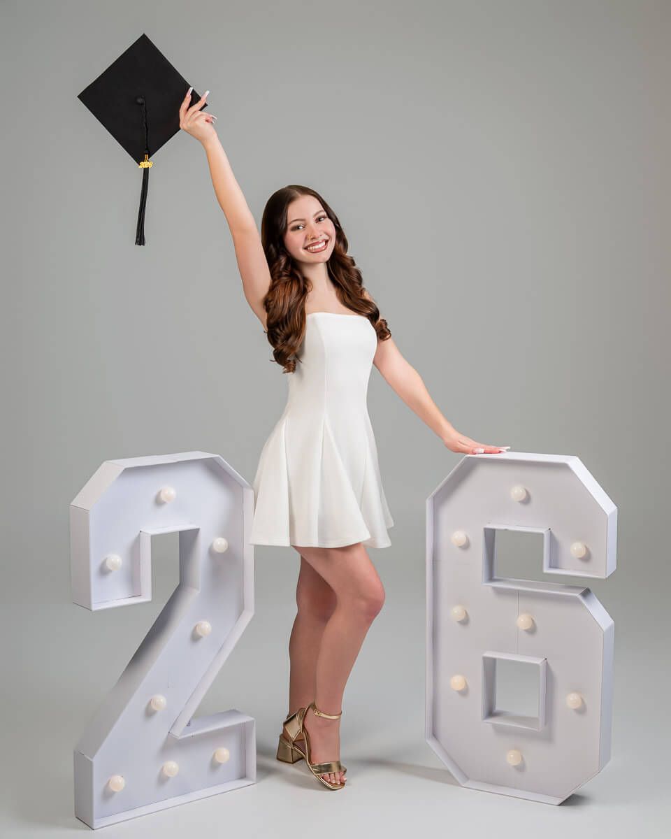 Smiling graduate in white dress holds cap aloft beside illuminated marquee numbers 26 in studio photo.