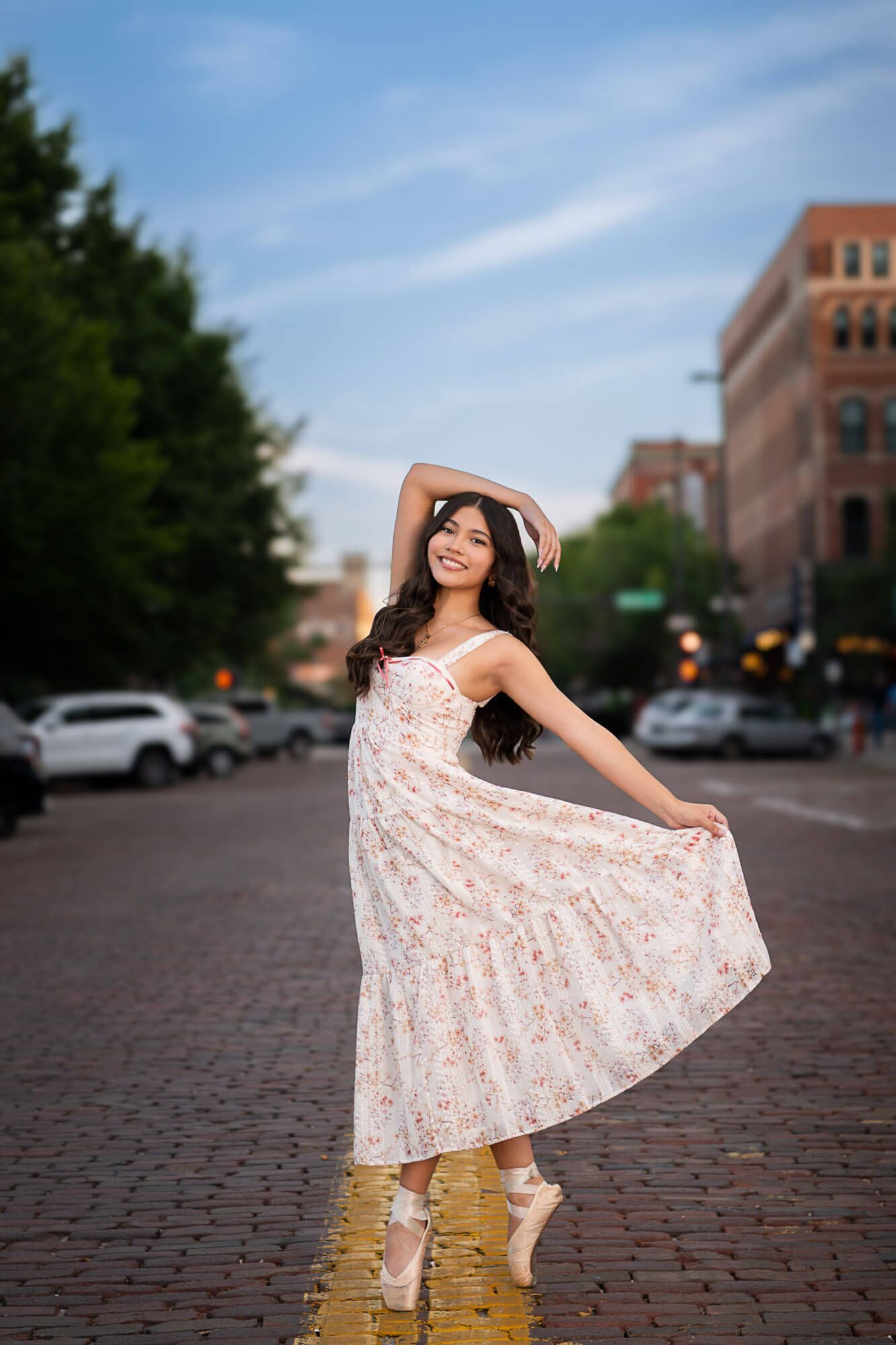 Playful poses in a floral dress on brick pavement with urban architecture in the background.