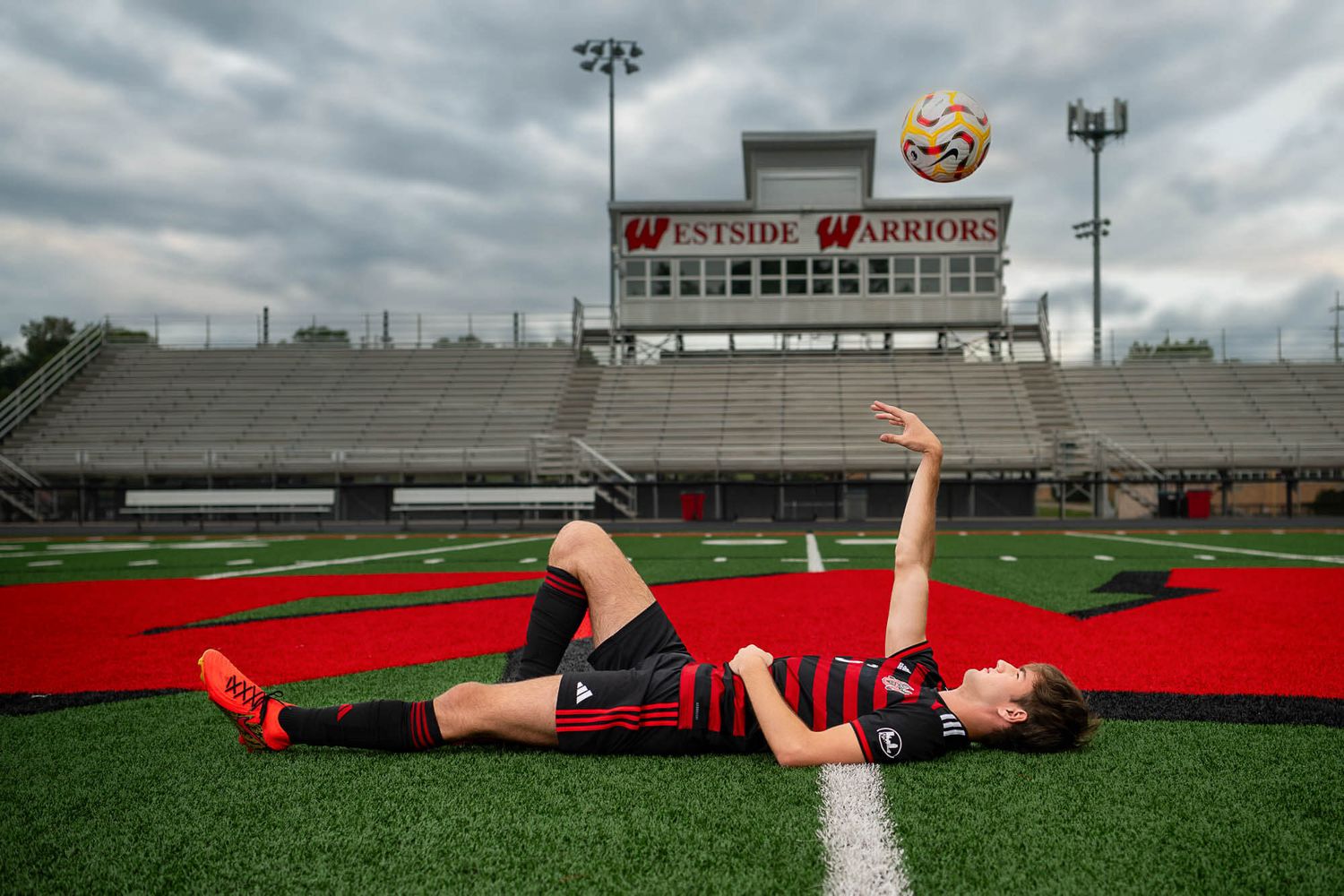 A person lies on a red turf football field beneath stadium bleachers and scoreboard on an overcast day.