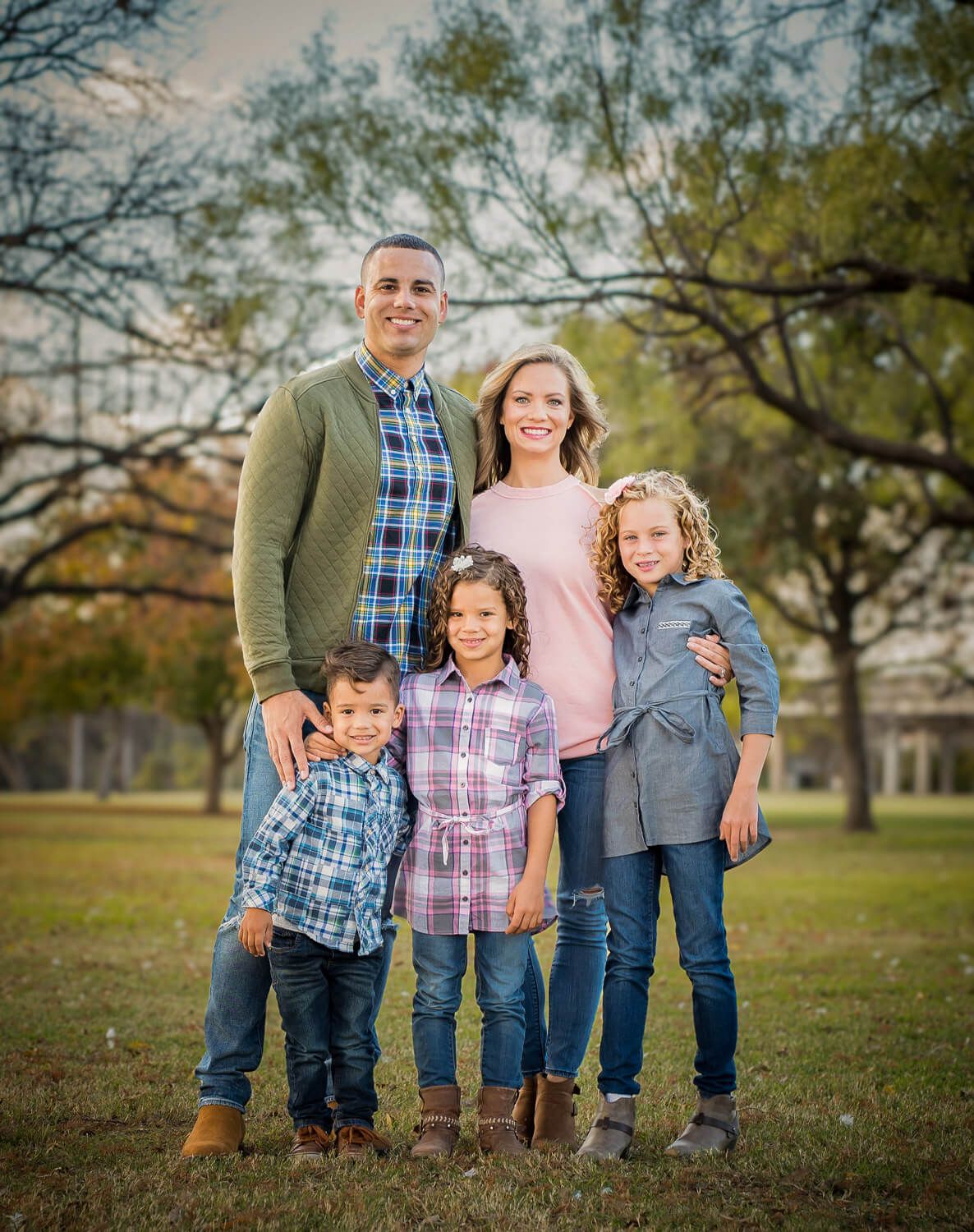 Family wearing coordinated blue and pink outfits poses together outdoors in a park setting during autumn.