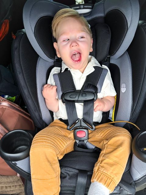 A child sits secured in a gray car seat wearing a white shirt and tan pants while making an excited expression.