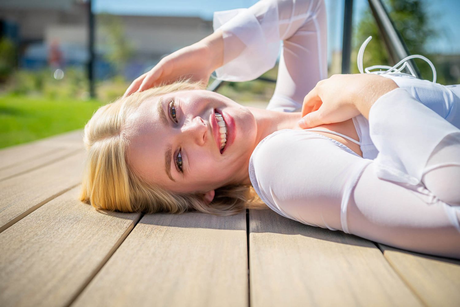 Smiling blonde woman in white top lying on wooden deck outdoors.