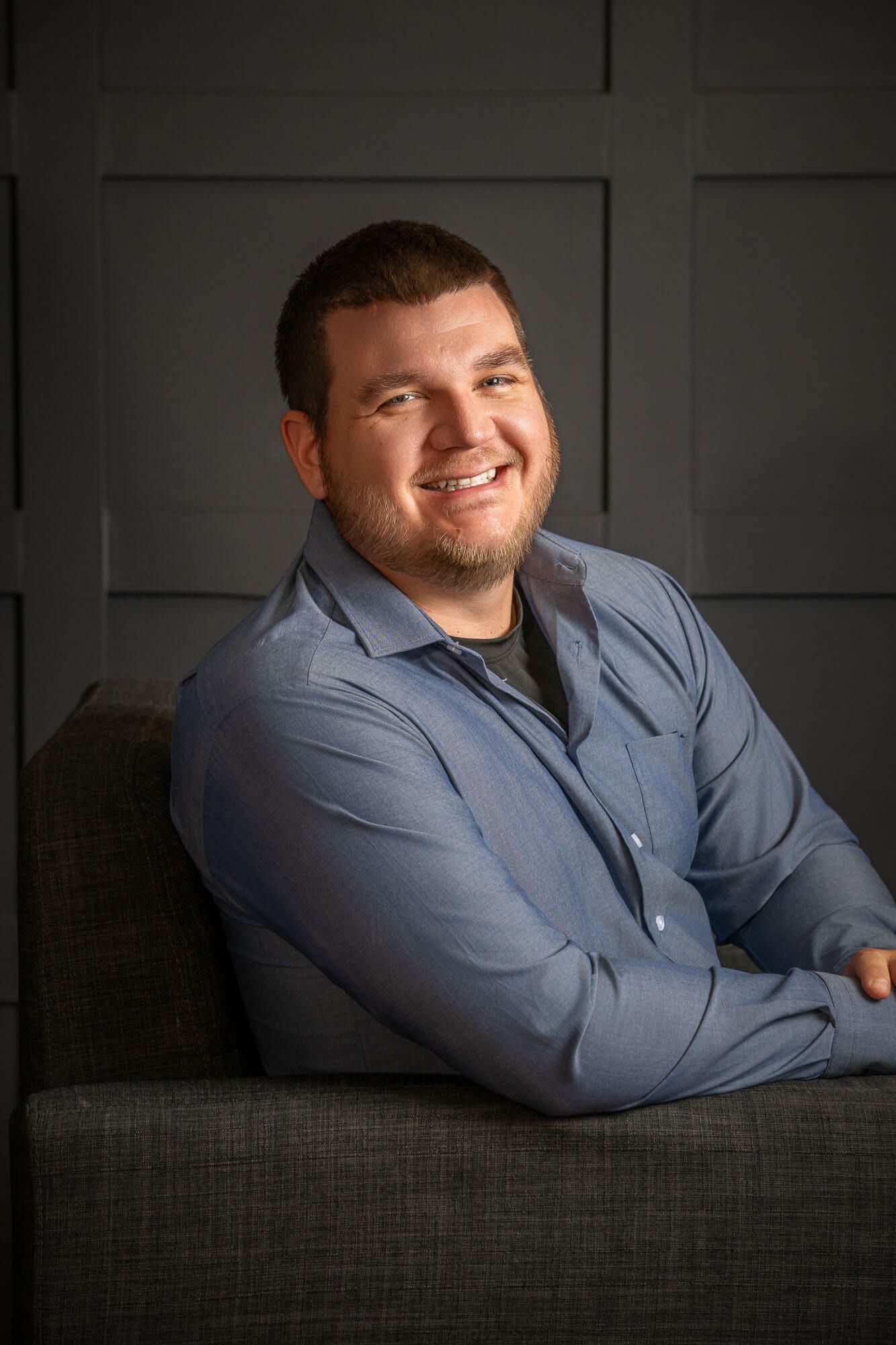 Casual business portraits of person reclining on grey couch in blue shirt.