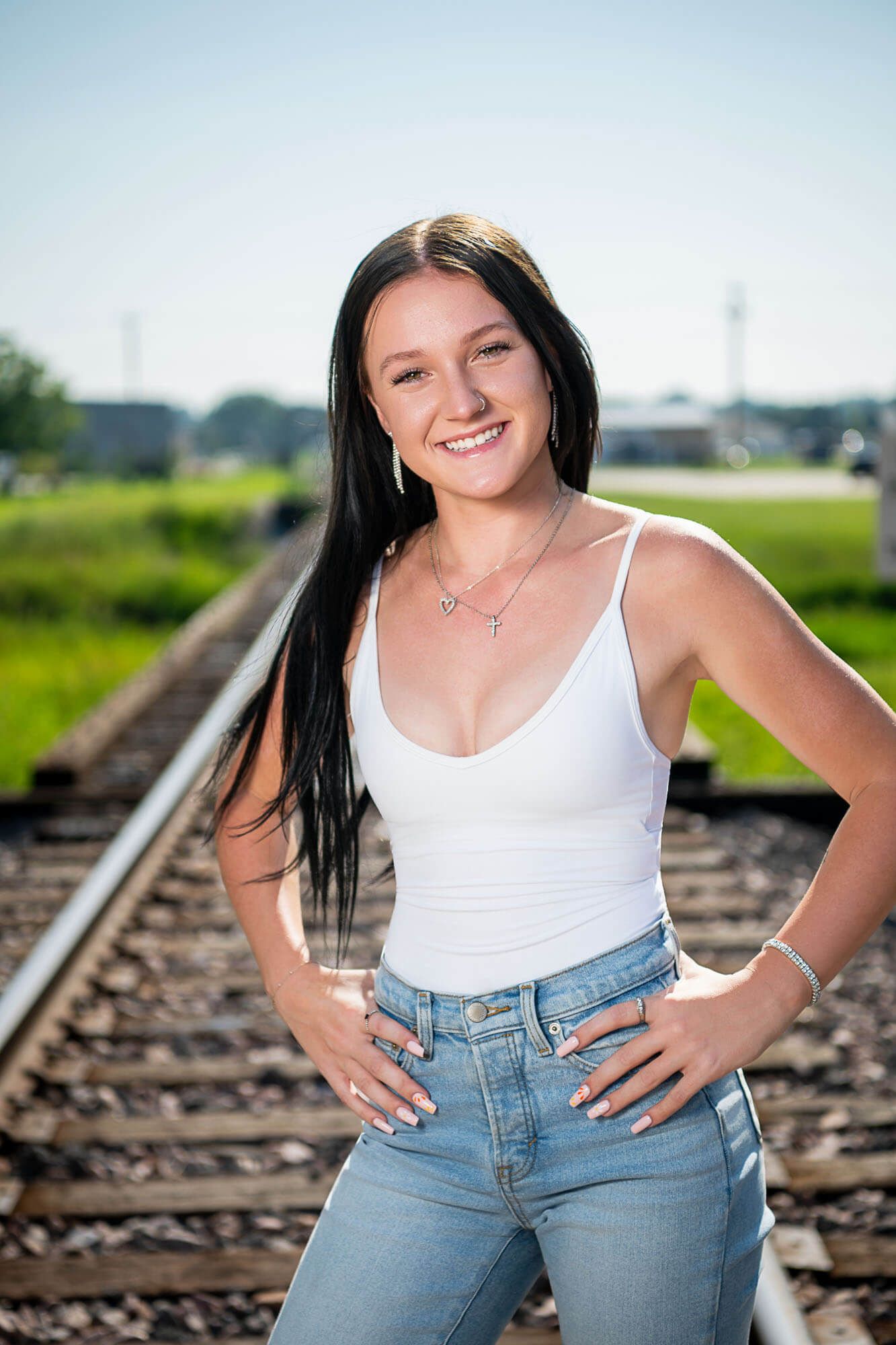 Person in white tank top and jeans poses casually on railroad tracks in outdoor portrait series.