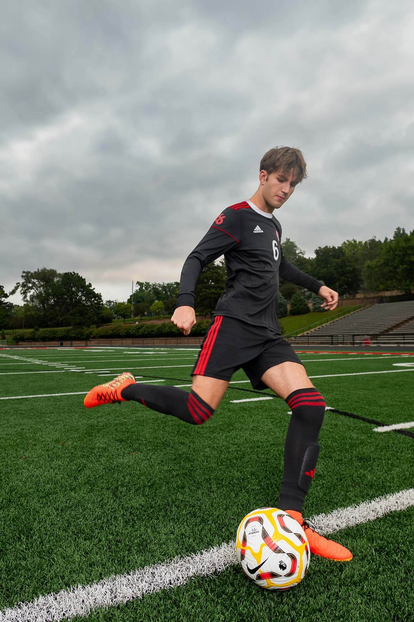 Soccer player in black uniform practices kicking techniques with multiple balls on artificial turf field under cloudy sky.