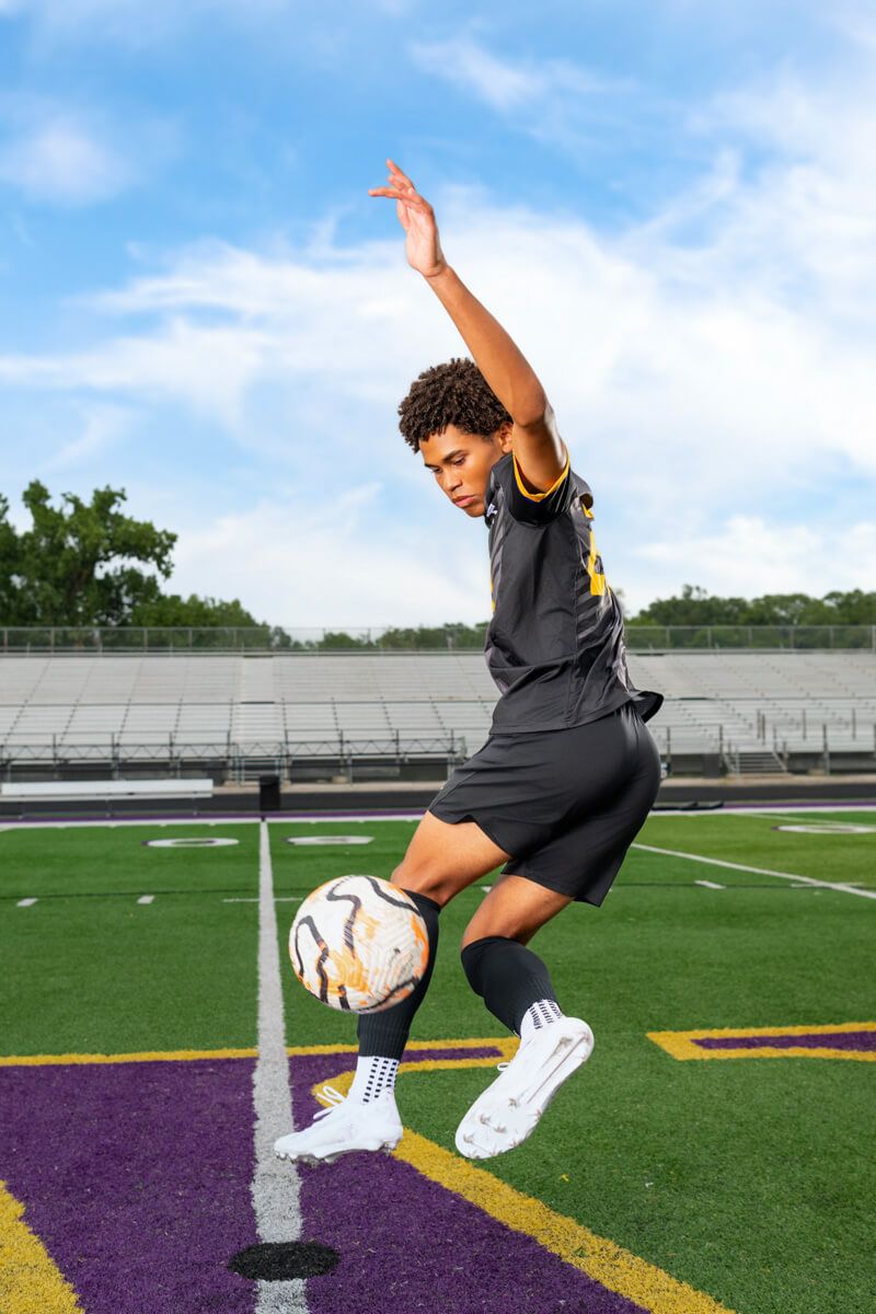Senior soccer player demonstrates kicking technique on athletic field with bleachers & purple endzone markings at Bellevue West High School.