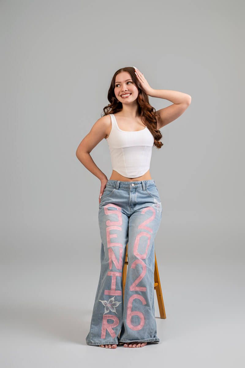 Smiling senior girl in white crop top and custom painted Senior 2026 flare jeans poses in studio.