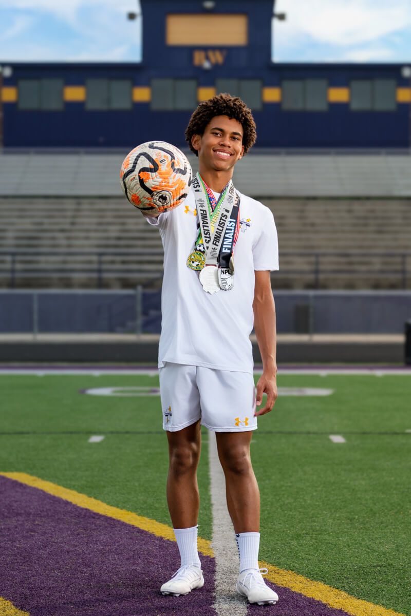 Young male soccer player in white uniform holds ball toward camera, wearing medals, standing on a stadium field.