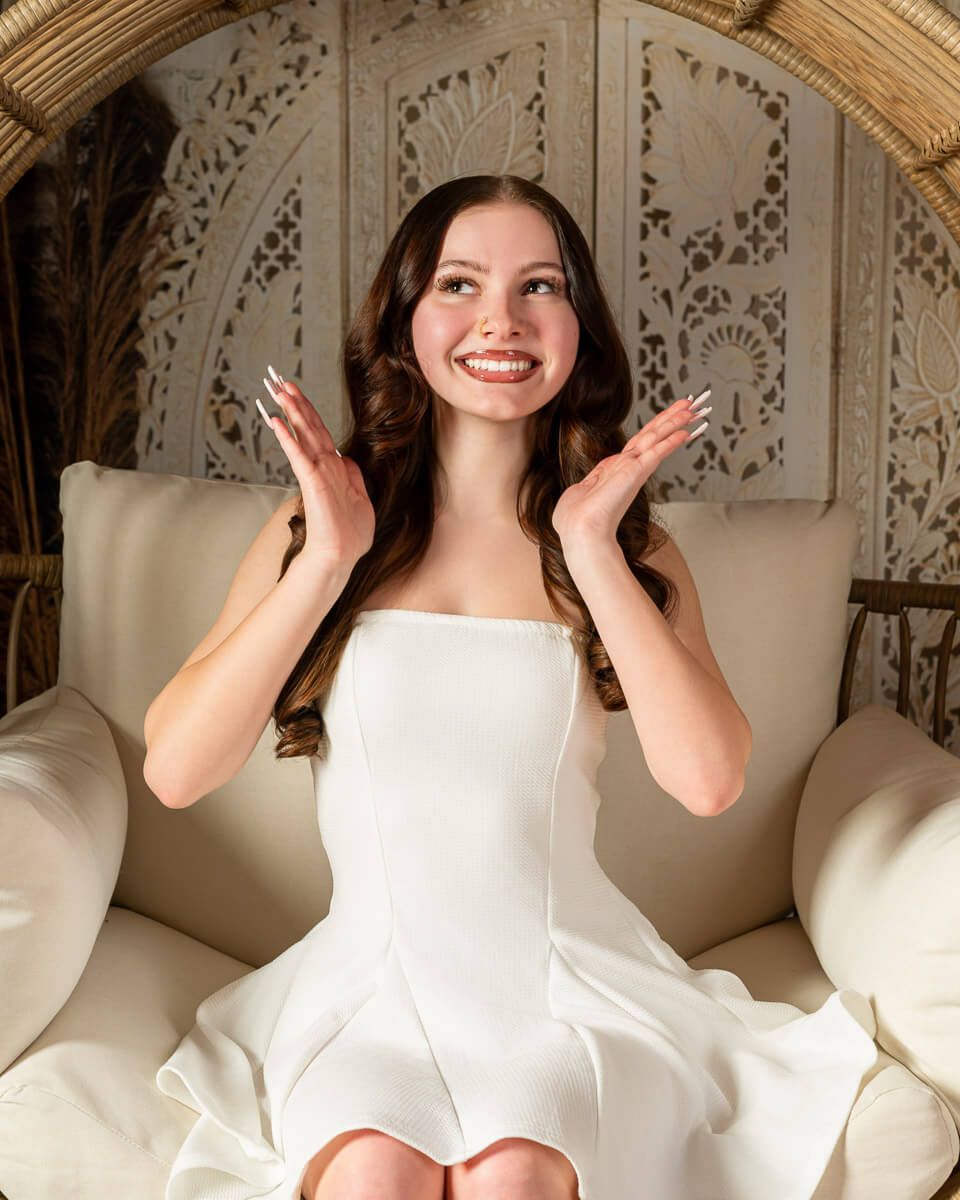 Smiling young woman in white strapless dress seated in wicker chair with hands raised expressively.