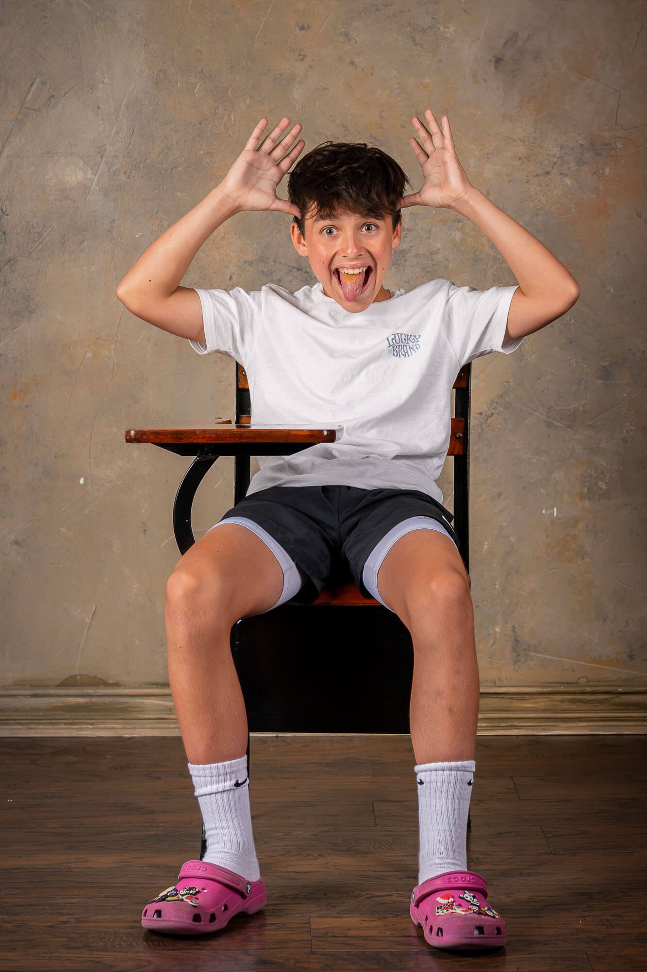 Energetic youth in white t-shirt and shorts sits in classroom chair wearing pink crocs and making playful expression.