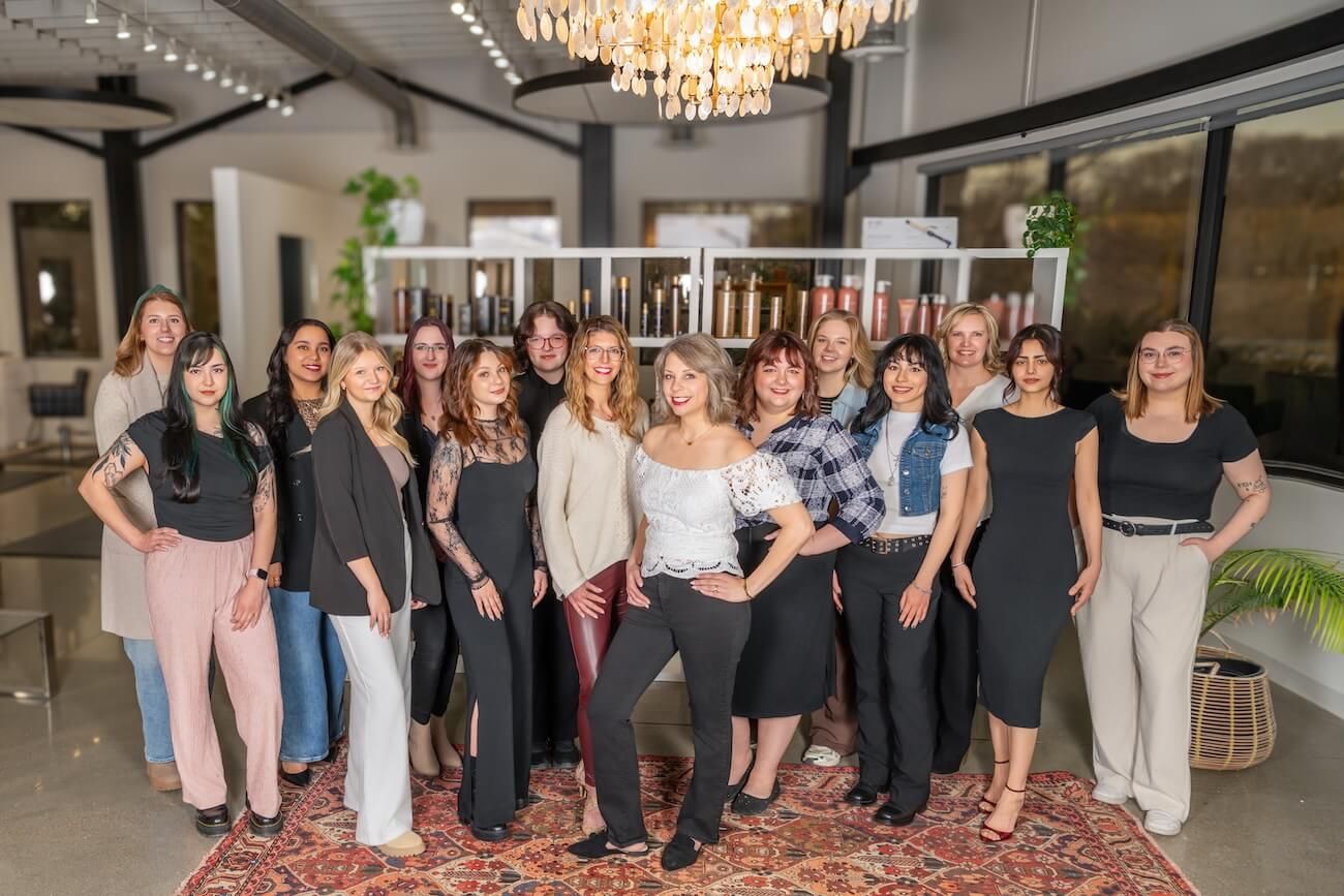 A group of women in business casual attire stand together smiling under a crystal chandelier in an elegant room.