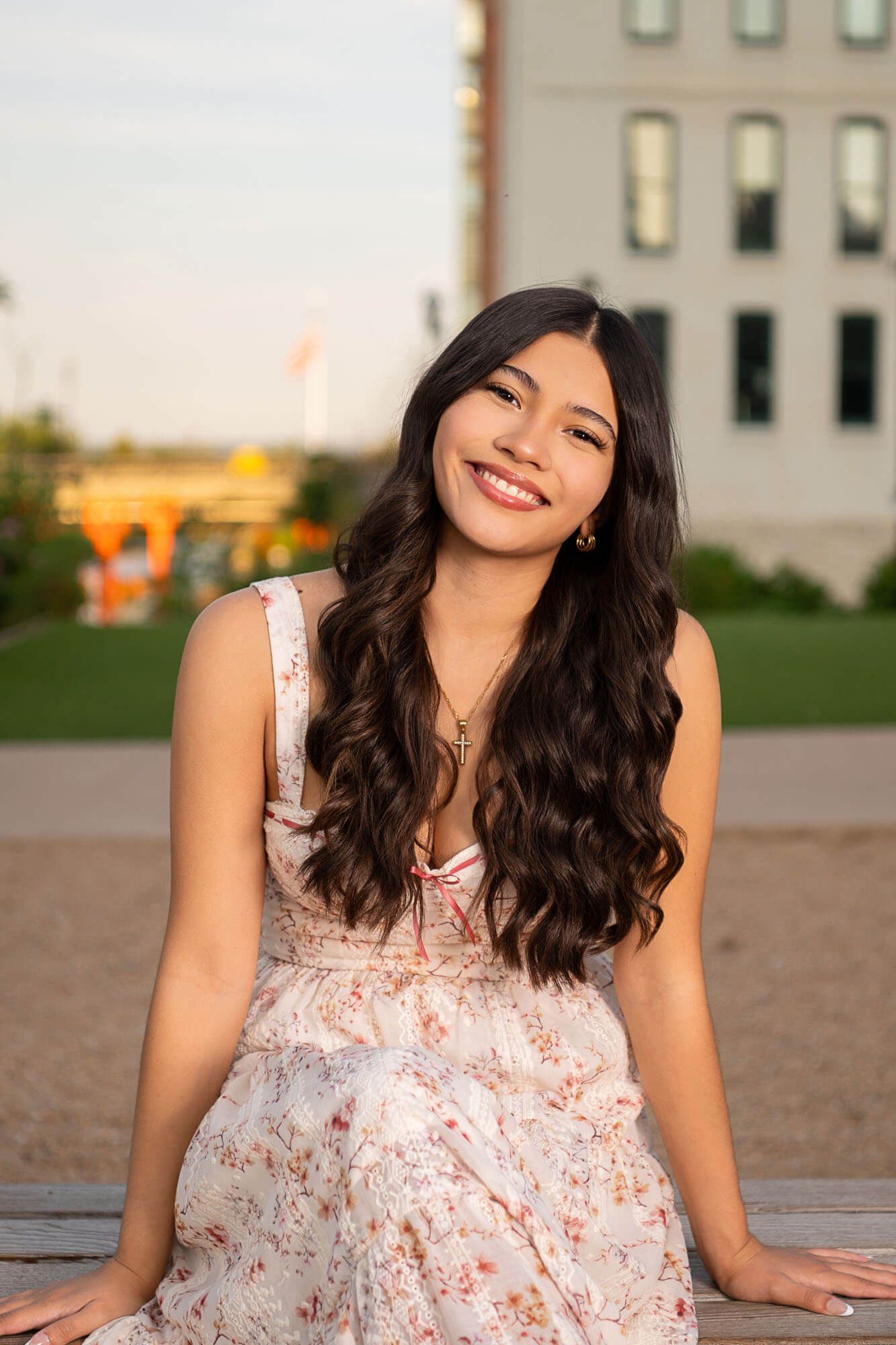 A person in a floral dress sits outdoors at sunset with a bright smile and long dark wavy hair.