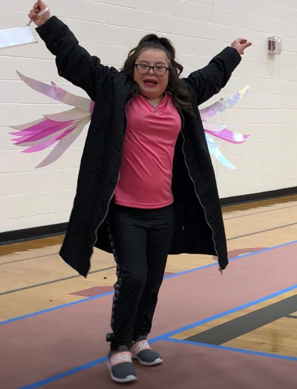 A person wearing a black jacket and pink shirt jumps joyfully with colorful paper wings in a school gymnasium.