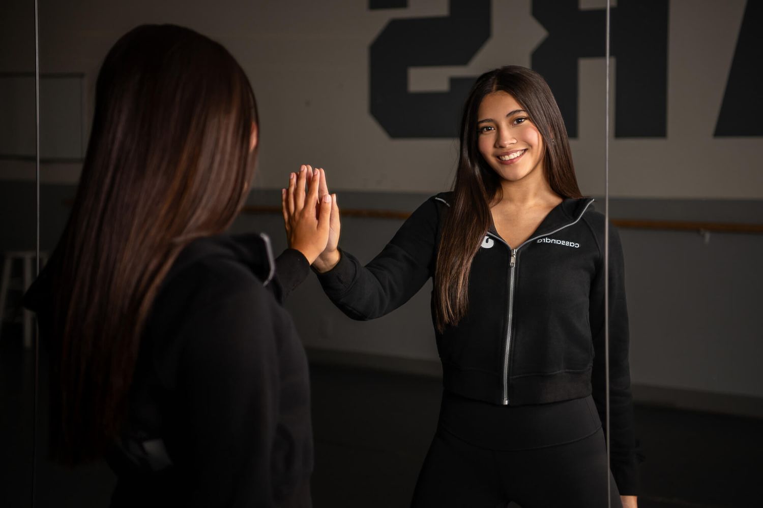 Woman in black zip-up jacket smiling at her reflection in a mirror during a workout session.