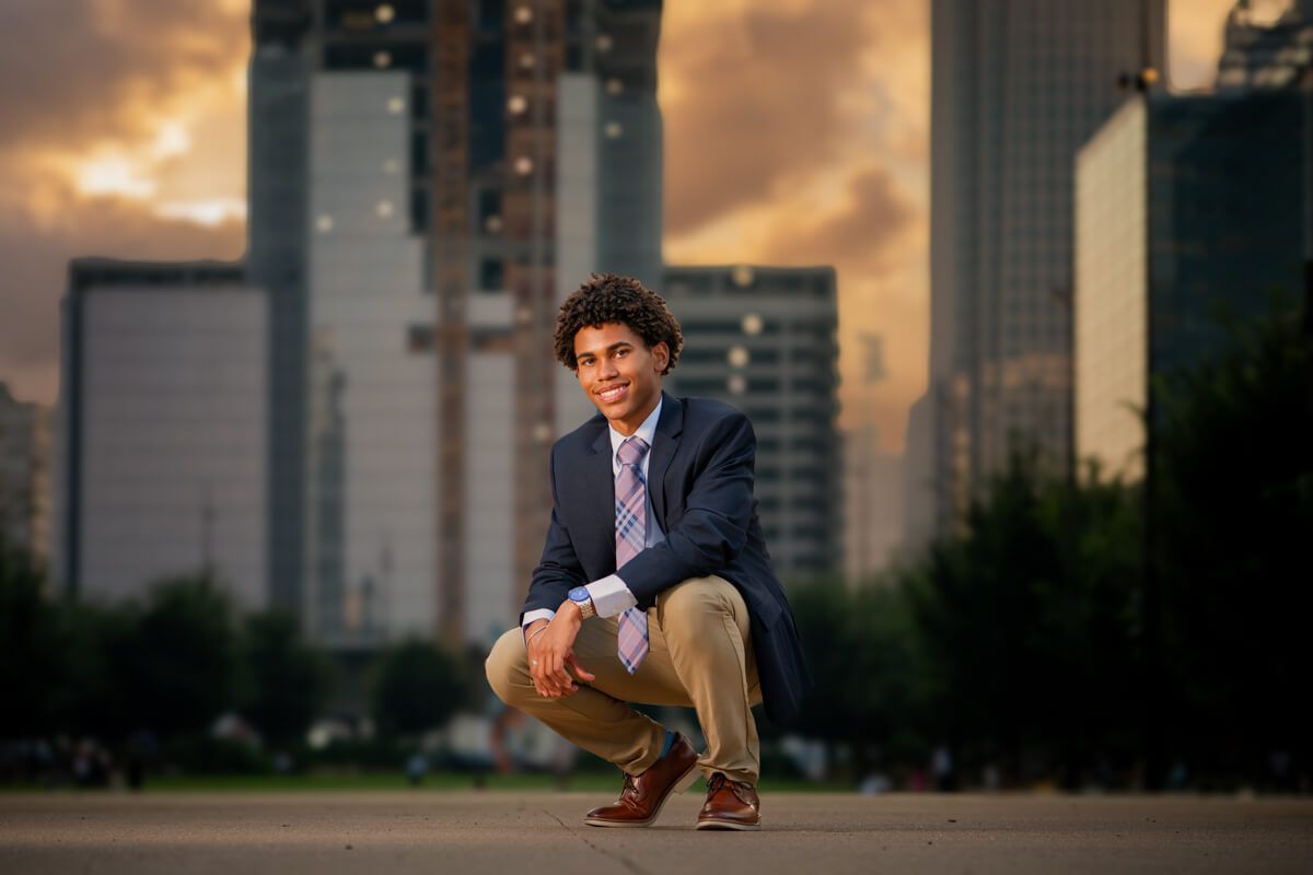Young man in blazer crouching in urban park with dramatic city skyline at sunset.