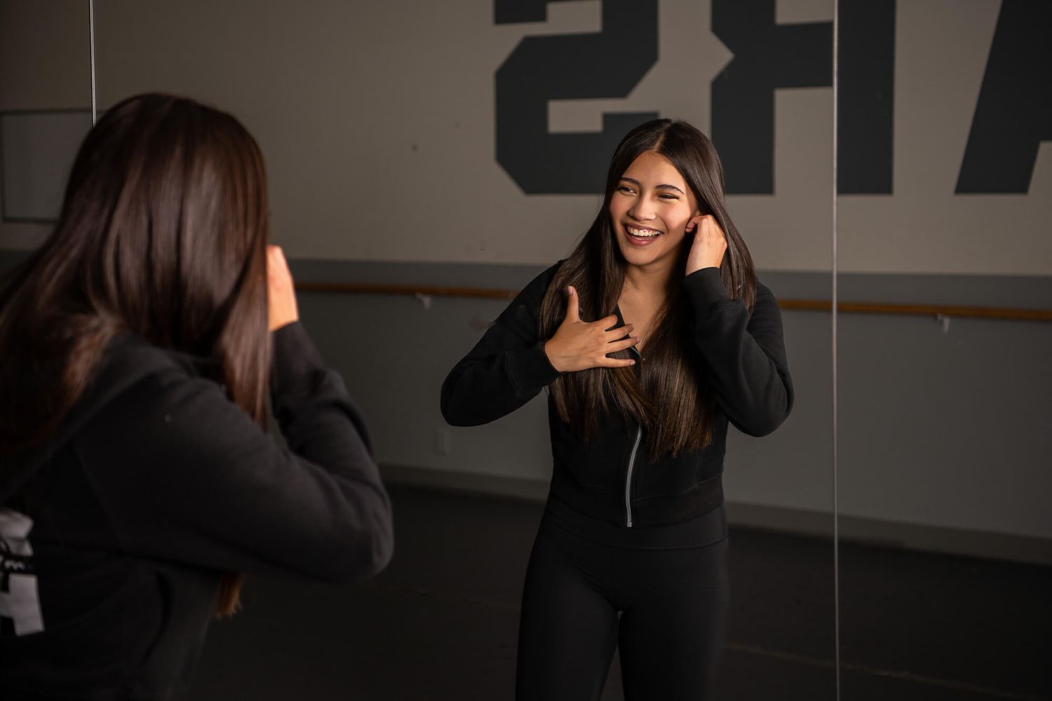A group of dancers in black attire practicing choreography in a dance studio with mirrors and ballet barre.
